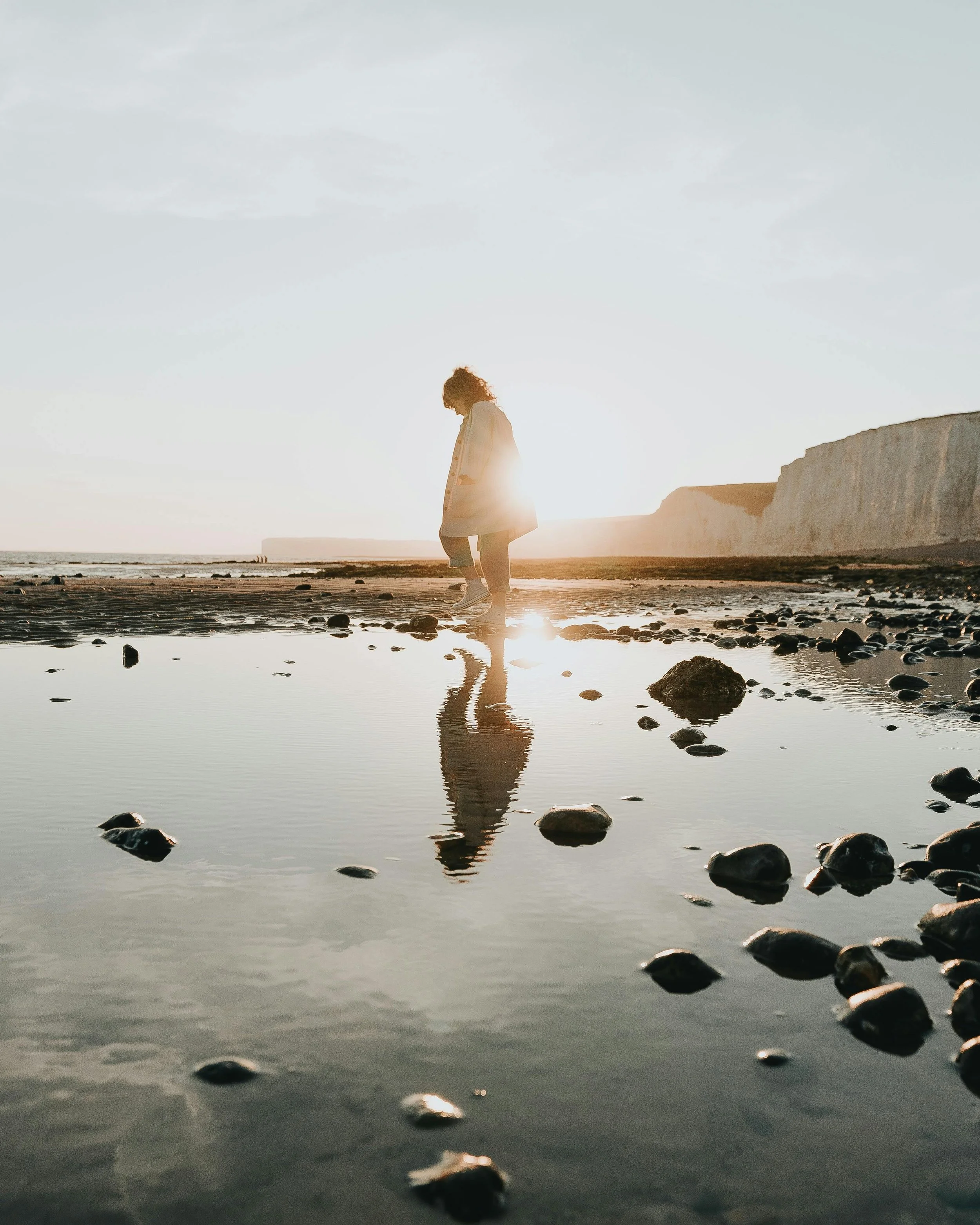 A woman stands on a rocky beach during sunset or sunrise, with her reflection visible in a small pool of water, and white cliffs in the background.