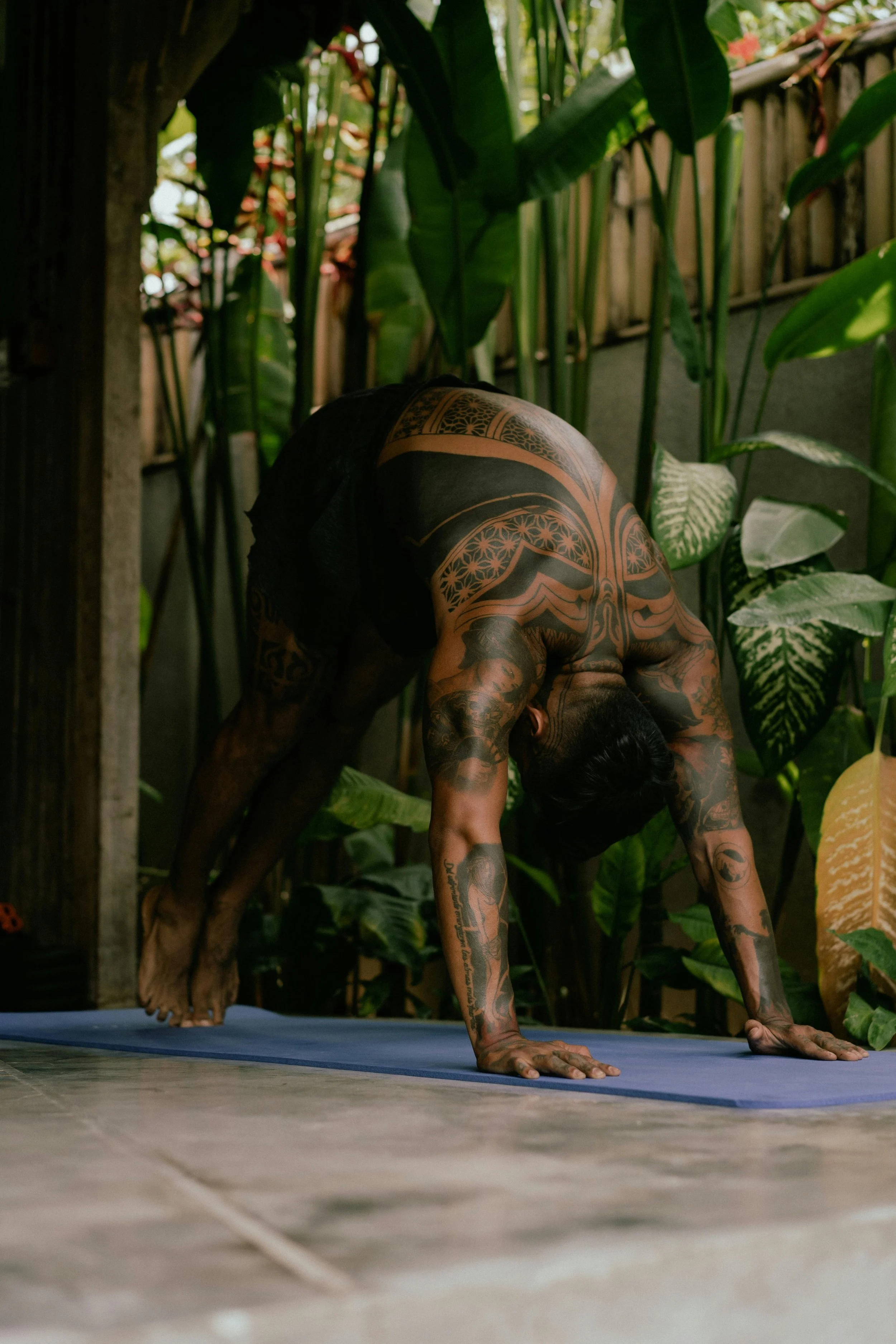 A tattooed man practicing yoga outdoors on a blue yoga mat, surrounded by green tropical plants.