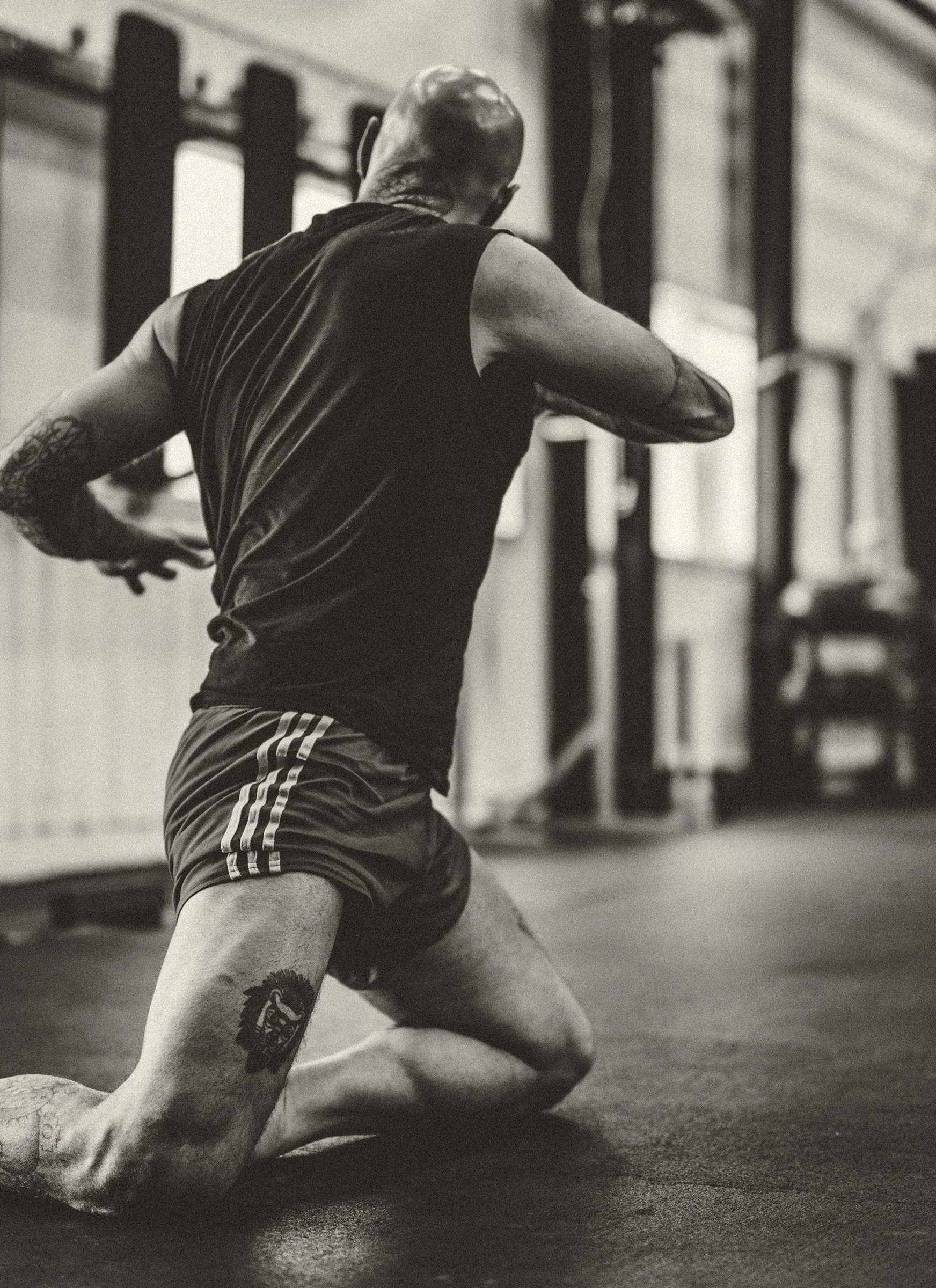A man with tattoos on his right arm and thigh, wearing athletic shorts and a sleeveless shirt, is kneeling on the gym floor in a crossfit gym, with his right knee on the ground and his left foot flat on the floor, engaged in a workout or stretching.