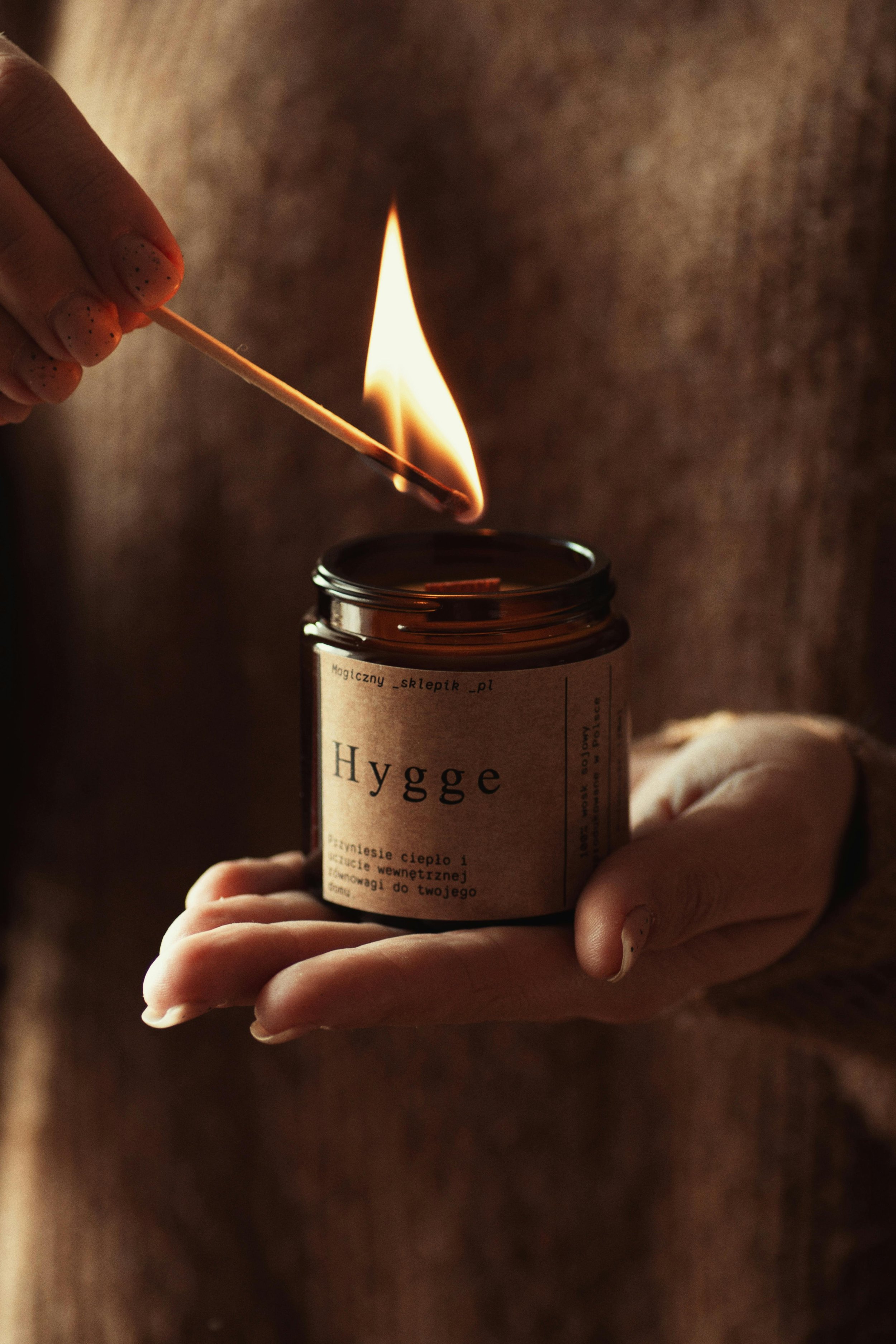 A person's hand holding a matchstick and a small jar labeled 'Hygge' with a lit matchstick about to ignite a candle or wick inside the jar.