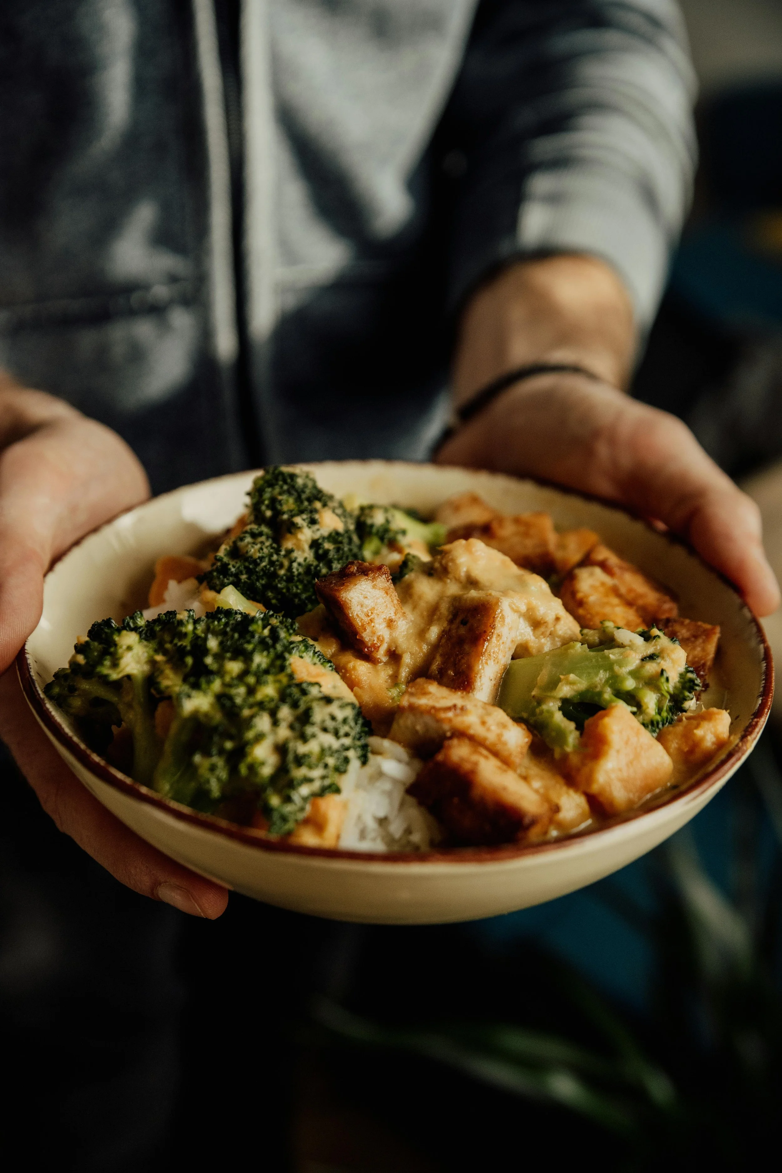 Person holding a bowl of rice with broccoli, chicken, and tofu.