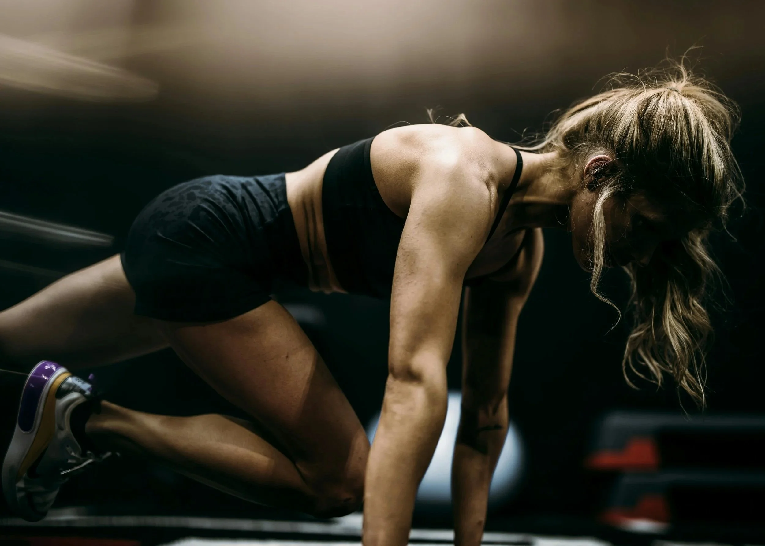 A woman in workout clothes is in a crouched position on a gym floor, likely doing a fitness exercise.
