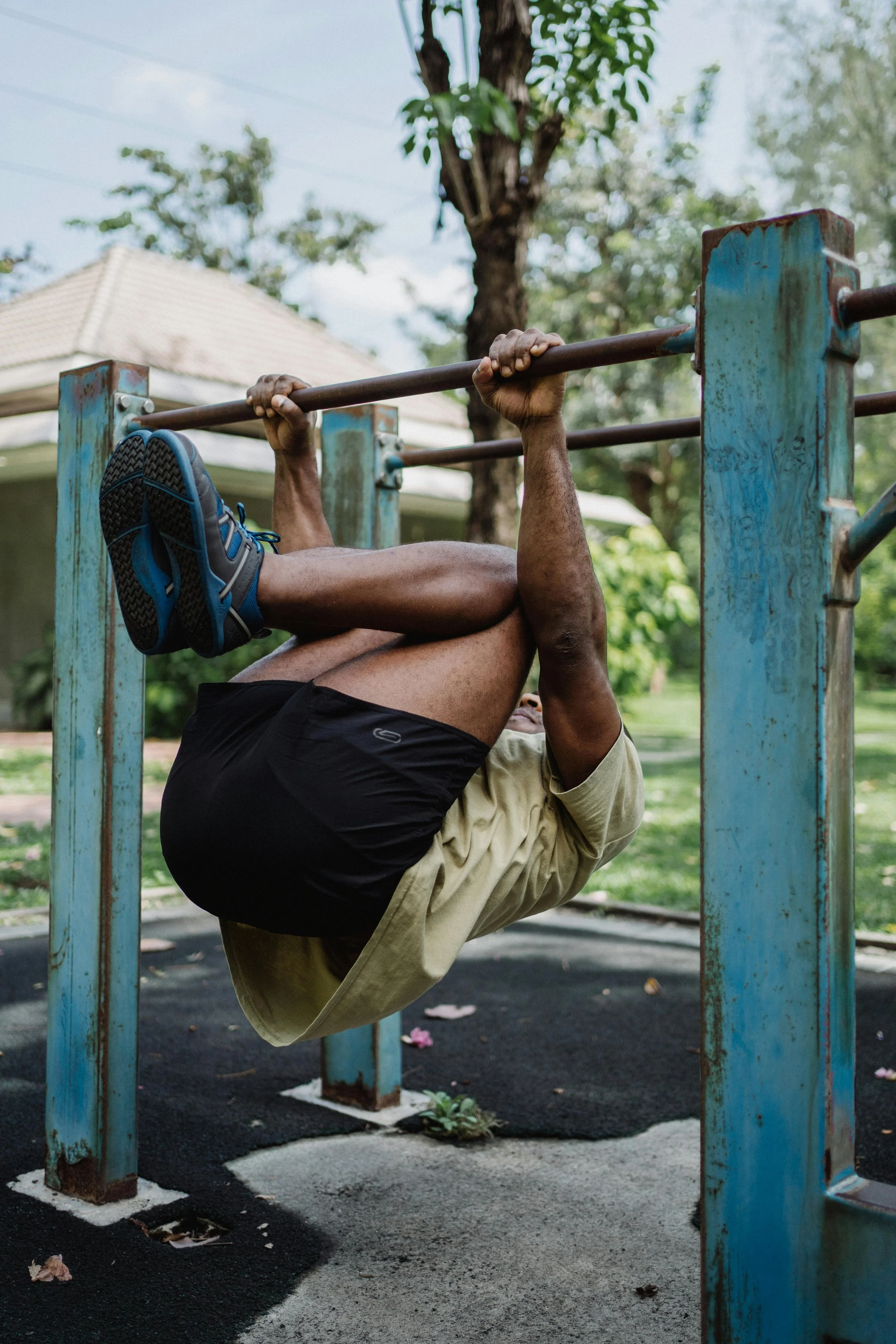 A man hanging upside down from a horizontal bar at a playground, with a beige shirt, black shorts, and blue shoes.