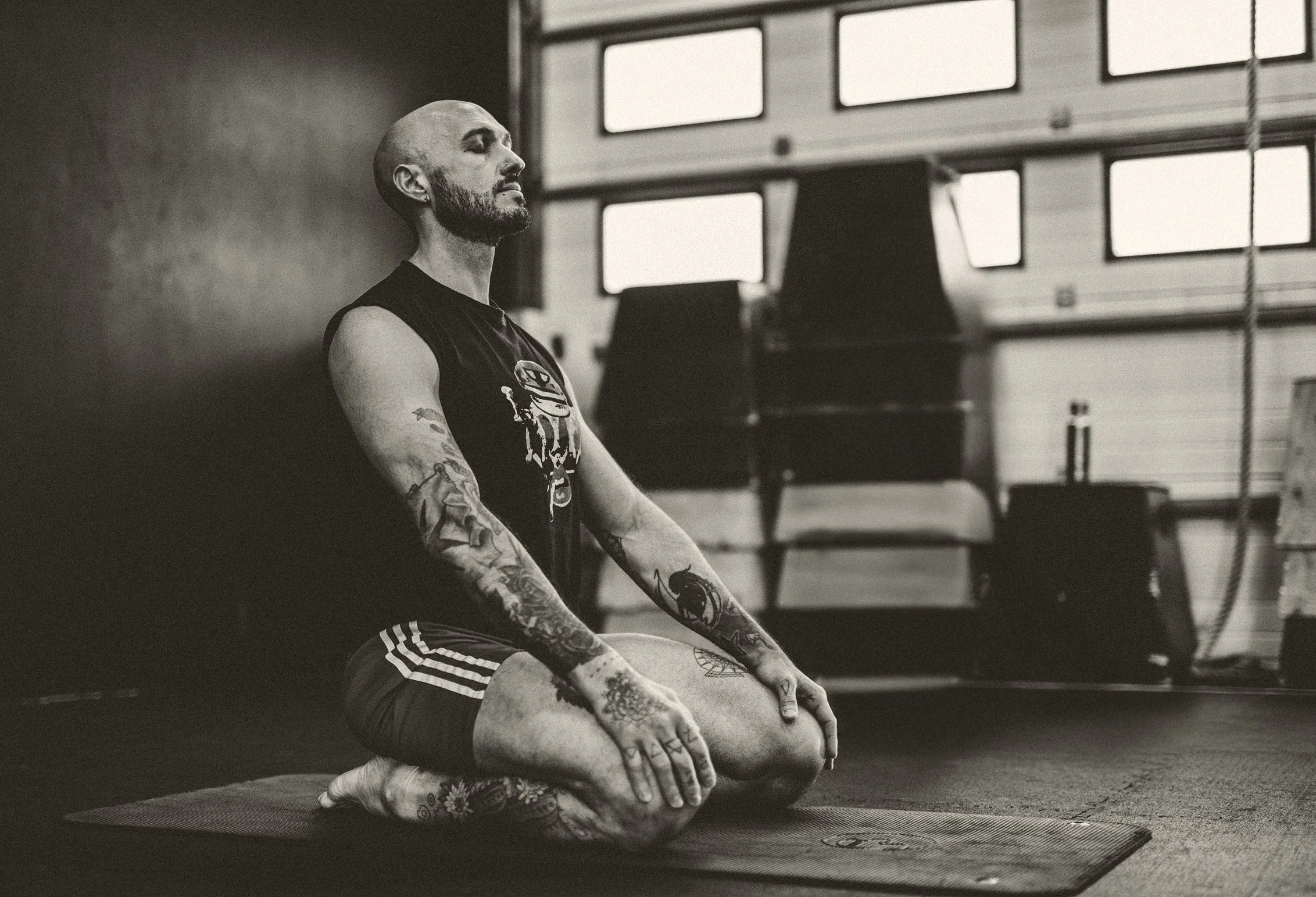 A man with tattoos practicing yoga in a garage, sitting in a cross-legged meditation pose on a mat with eyes closed.