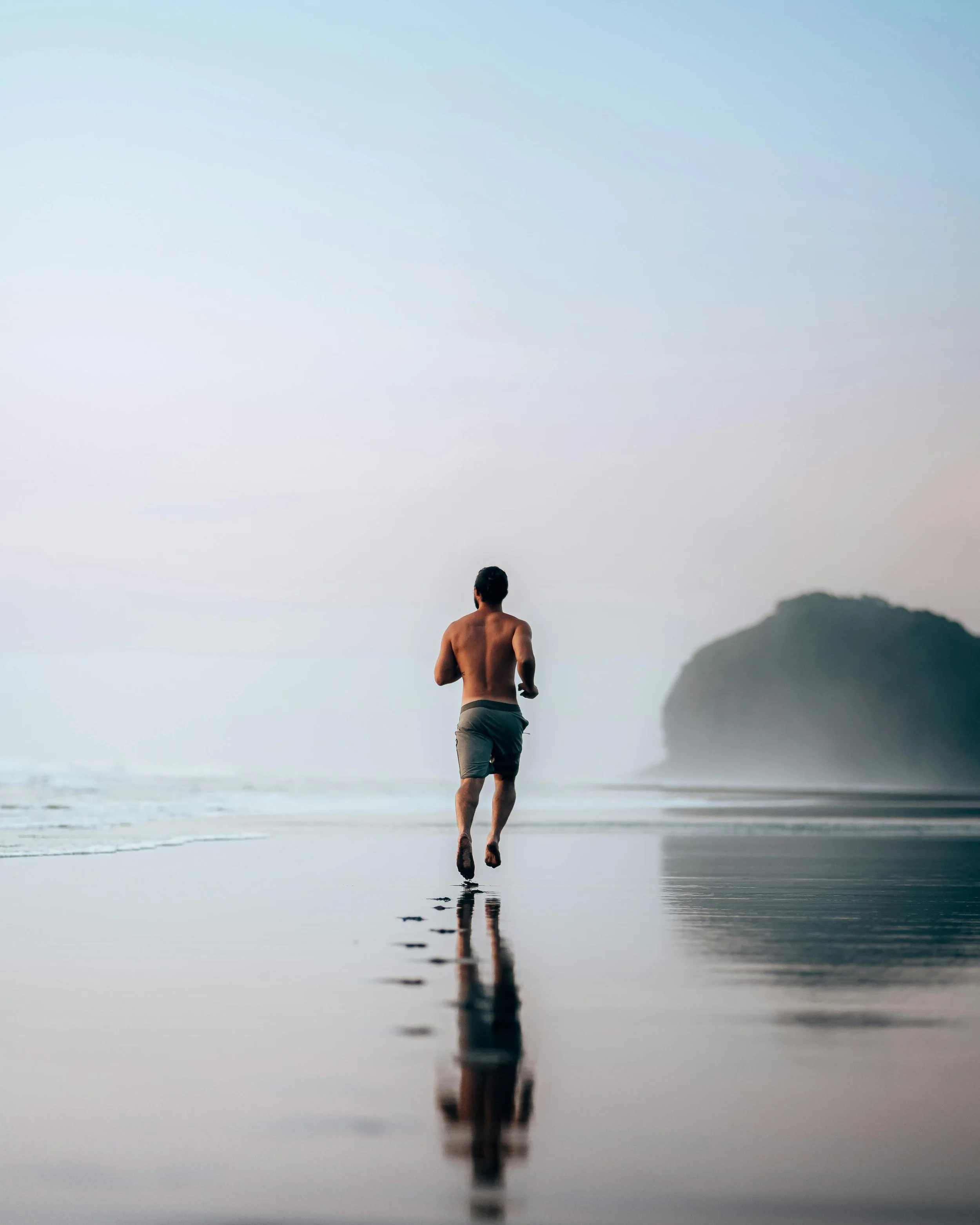 A man jogging on a wet beach shoreline during dusk or dawn, with a large island or rock formation in the distance and his reflection in the shallow water.