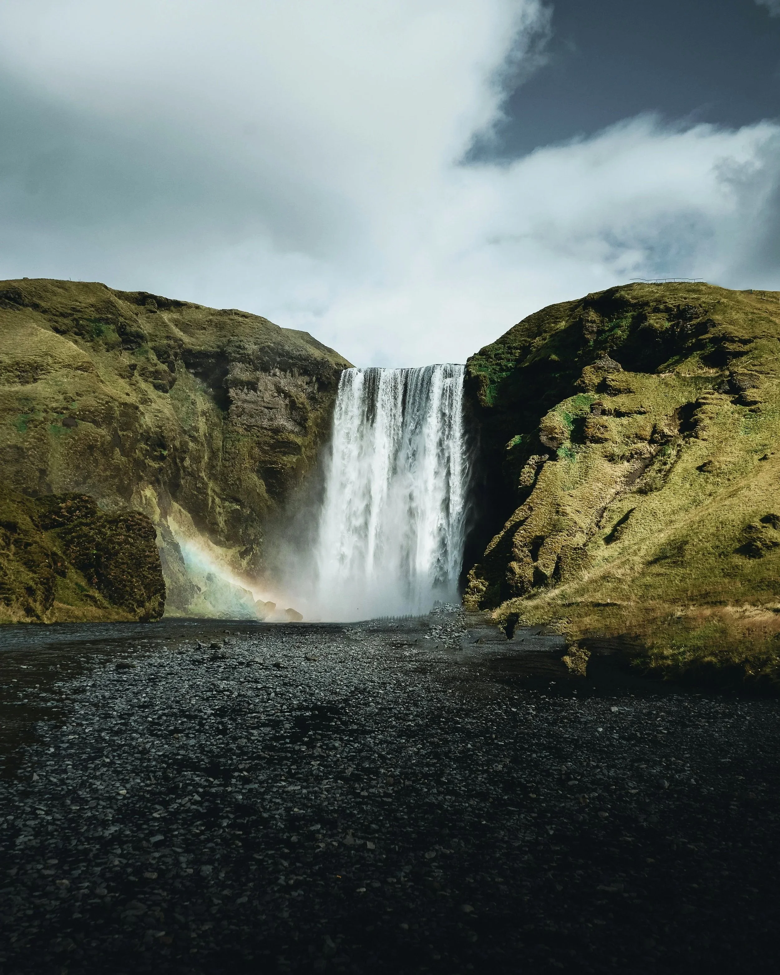 Waterfall cascading down a rocky cliff into a pool below, with cloudy sky overhead and a faint rainbow near the base.
