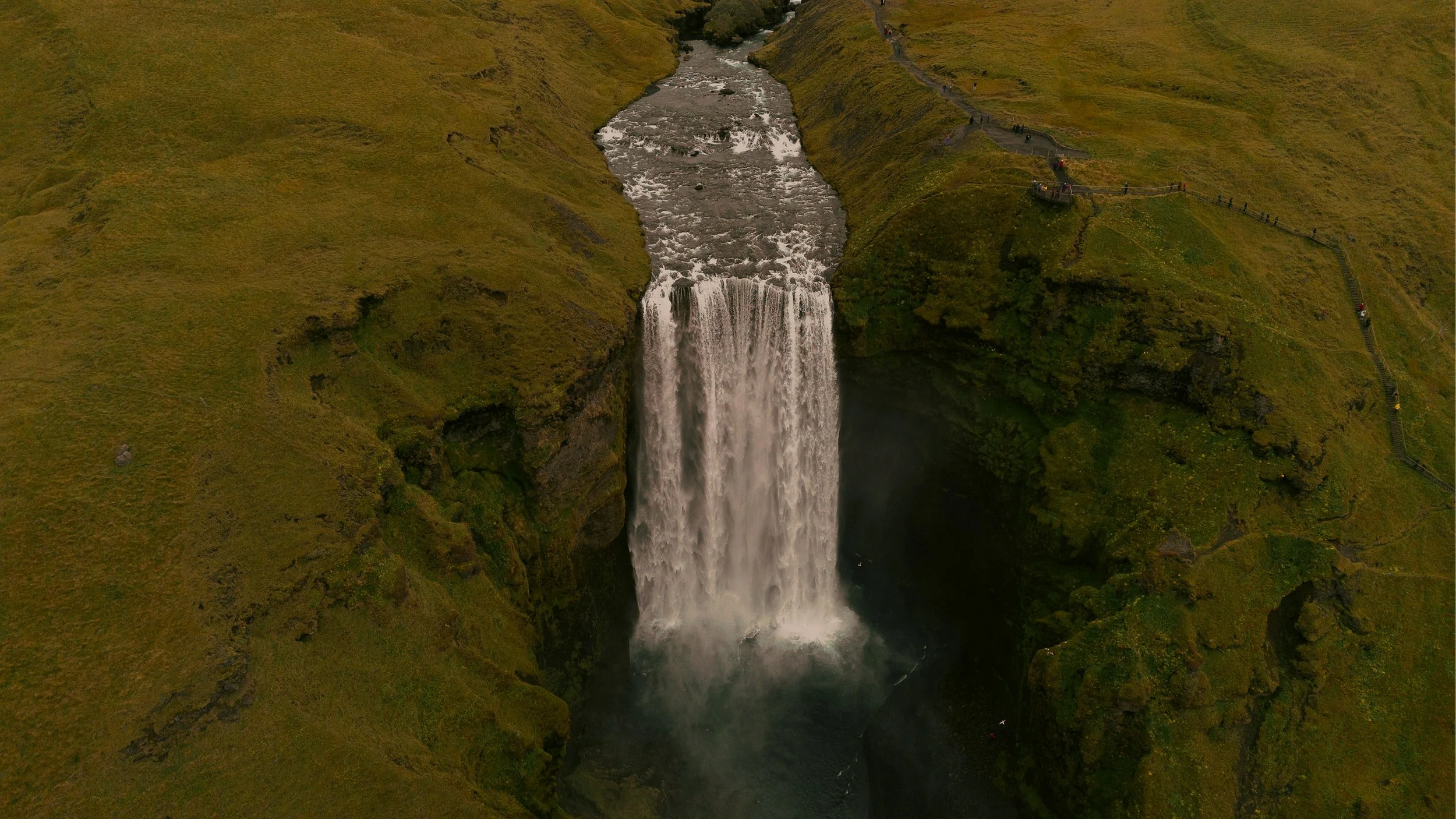 Aerial view of a waterfall flowing over a cliff, surrounded by green grassy hills with pathways and visitors walking along the edges.