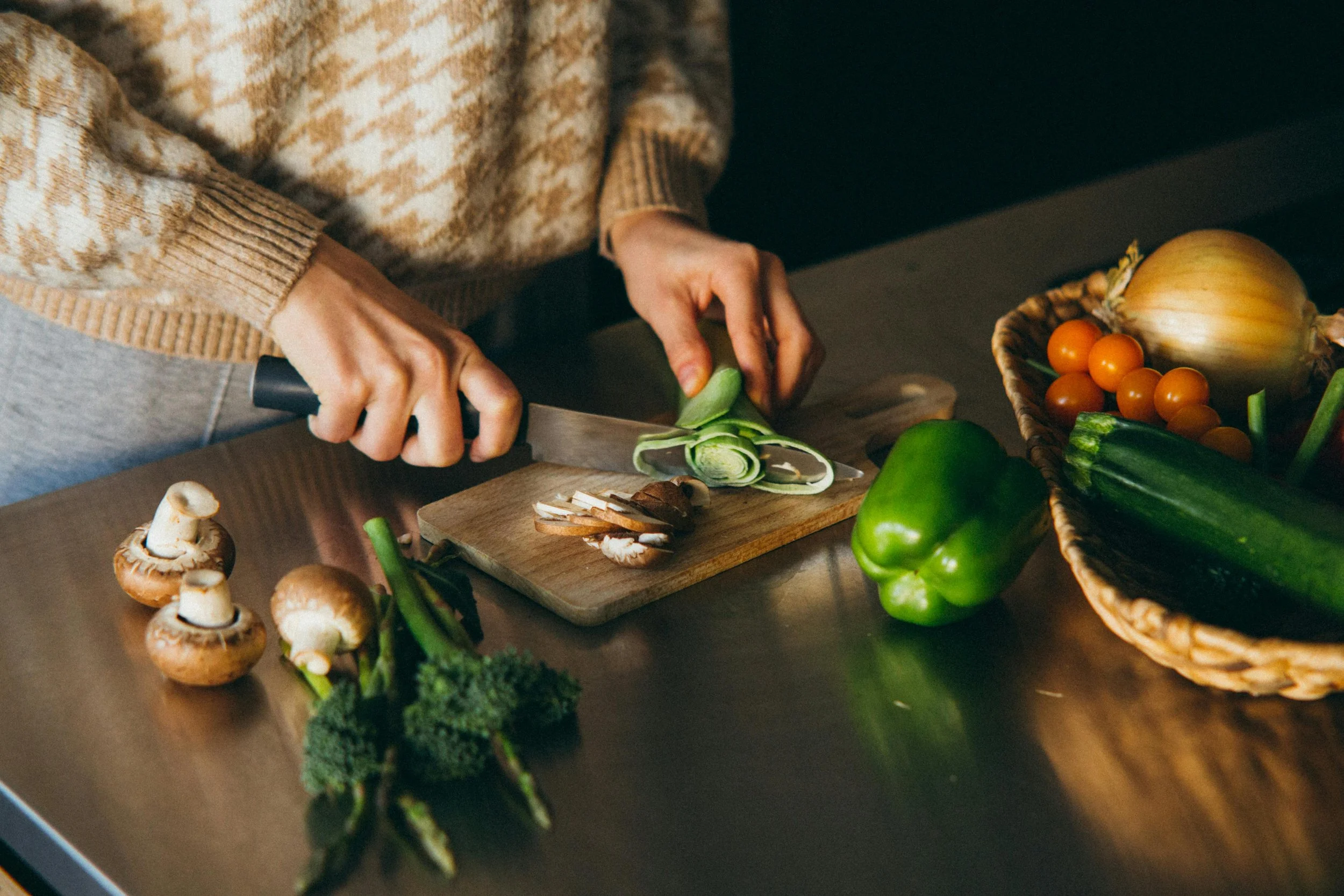 Person chopping green onions on a wooden cutting board, surrounded by various vegetables including mushrooms, broccoli, a green bell pepper, a cucumber, cherry tomatoes, and an onion, on a kitchen countertop.