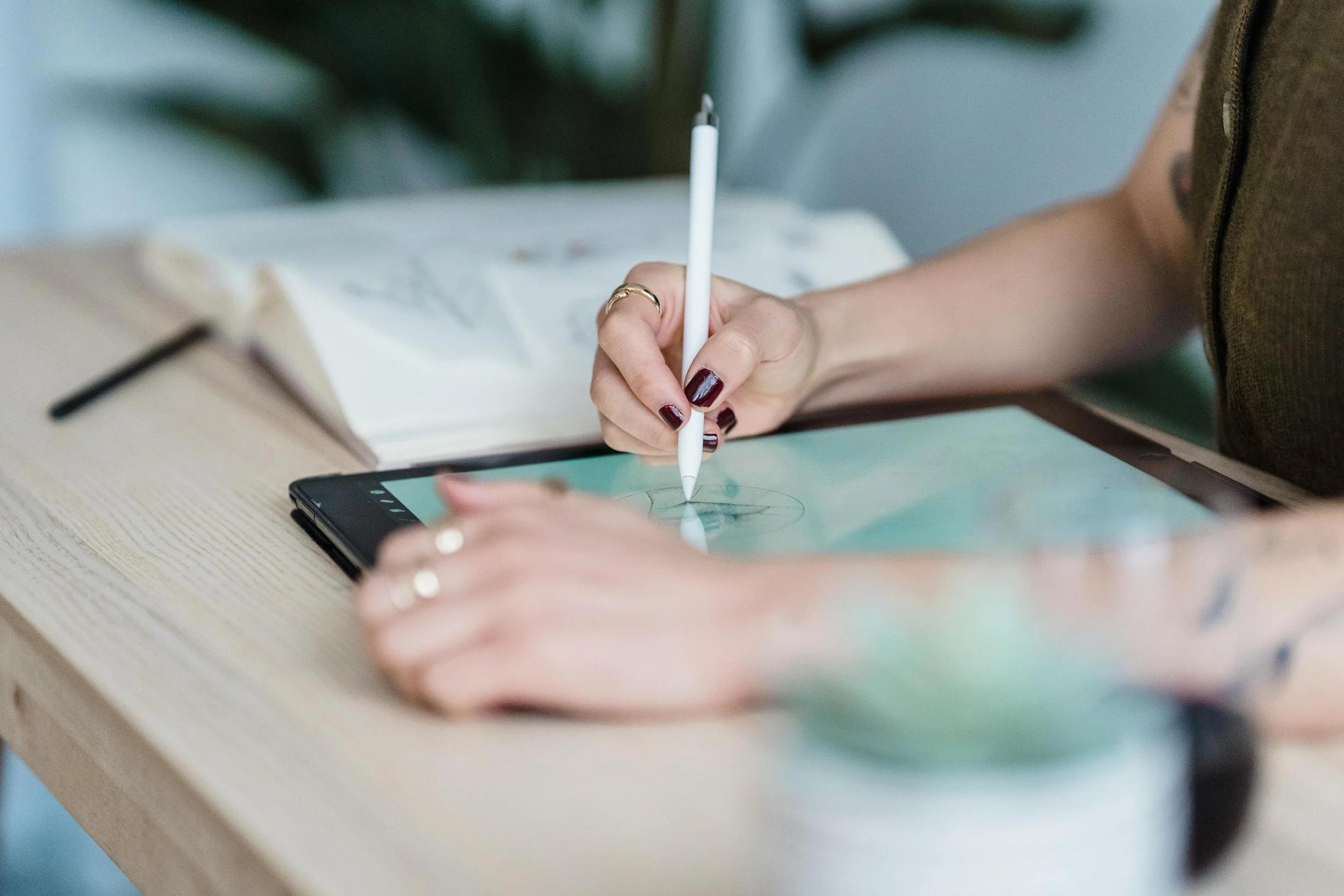 A woman using a stylus on a digital tablet, with a notebook and pen on the table.