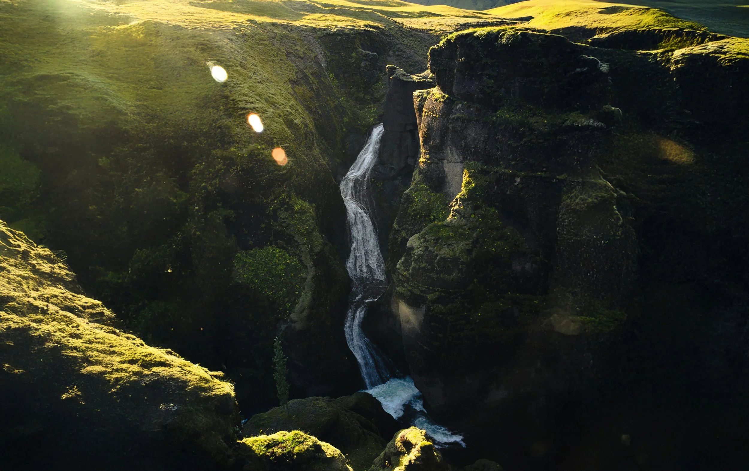 A small waterfall flowing between moss-covered rocks in a lush green canyon at sunset.