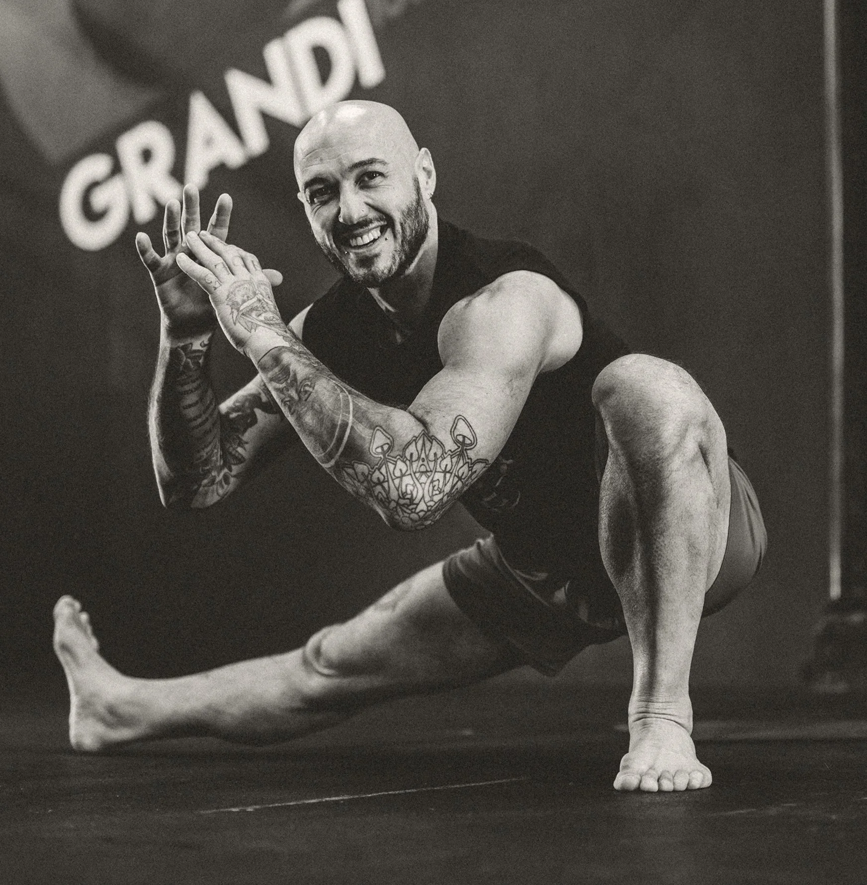A smiling muscular man with tattoos on his arms, bald head, and beard, performing a yoga pose on a gym floor. There is a sign in the background with the word 'GRAND'.