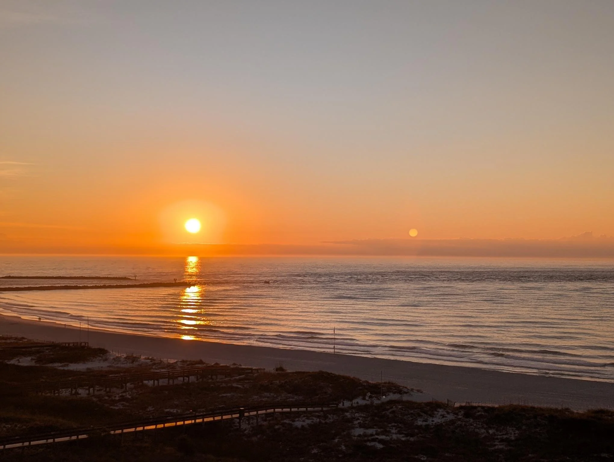 Sunset over the ocean with a calm, rippling sea and a sandy beach in the foreground.