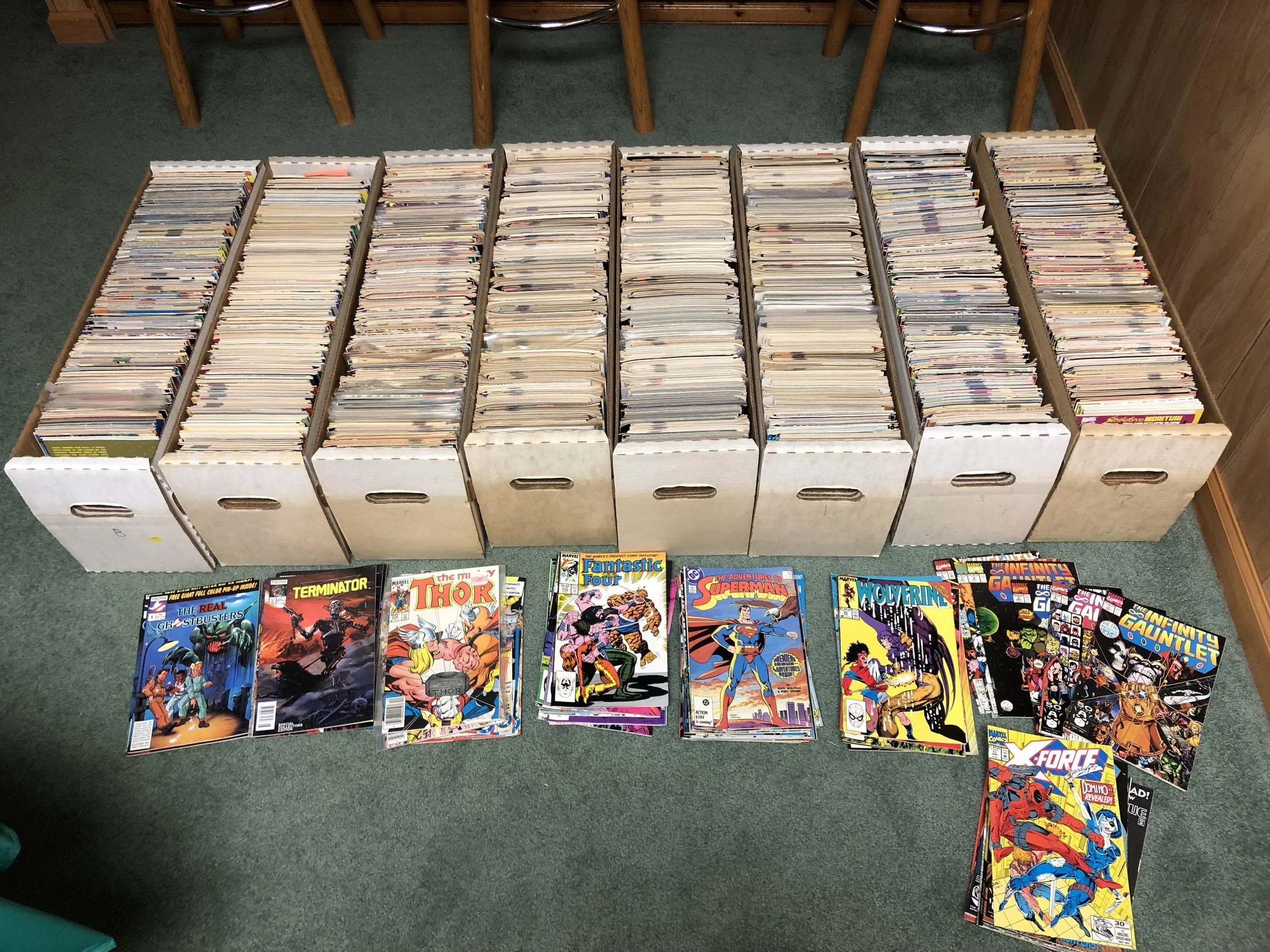 Collection of comic books in cardboard storage boxes and several on the floor, including titles like The Real Ghostbusters, Terminator, and Thor.
