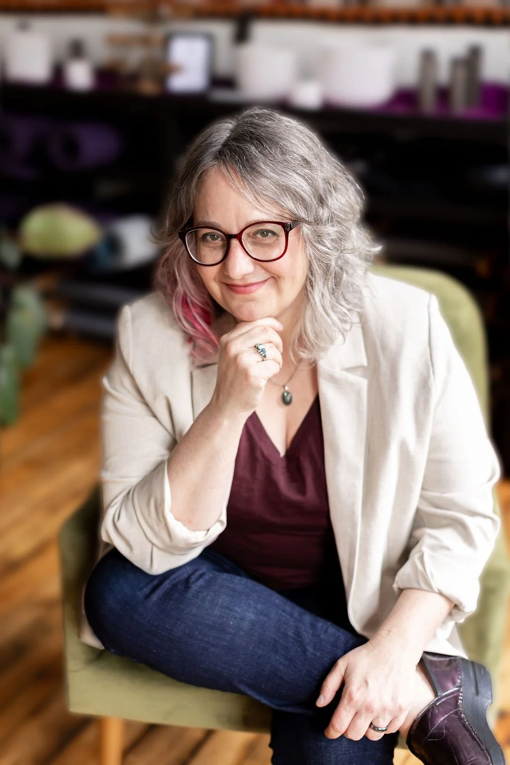 Portrait of a woman with curly gray hair and glasses, sitting on a green chair, smiling and resting her chin on her hand, in a cozy indoor setting.