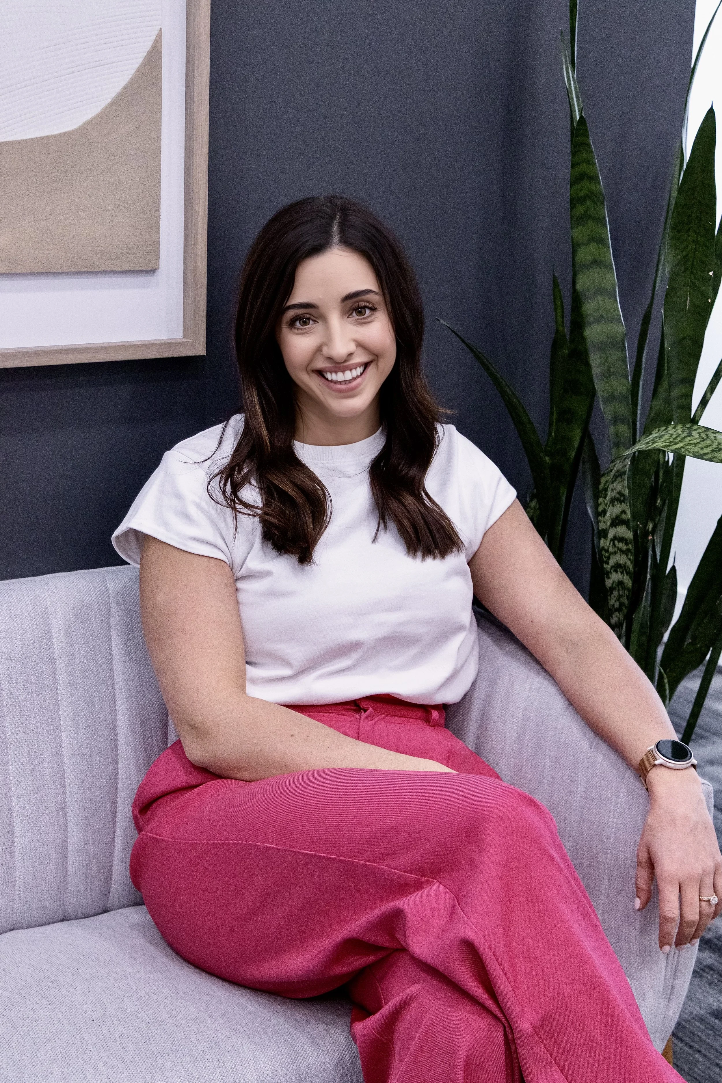 A woman with long dark hair, wearing a white t-shirt and pink pants, sitting on a gray couch in a modern living room with a dark wall, a plant, and abstract artwork.