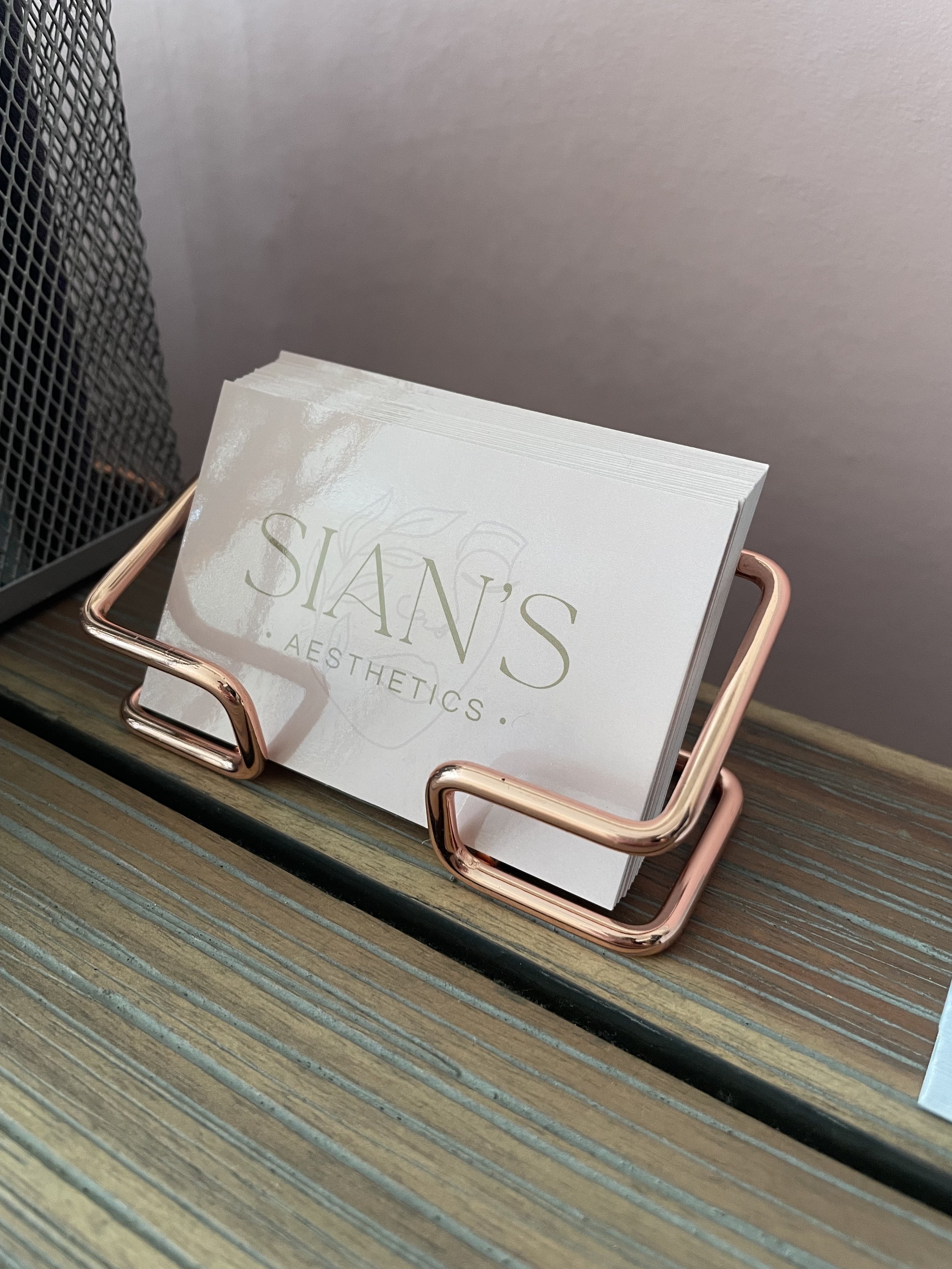 Business cards for "Sian's Aesthetics" in a rose gold holder on a wooden surface.