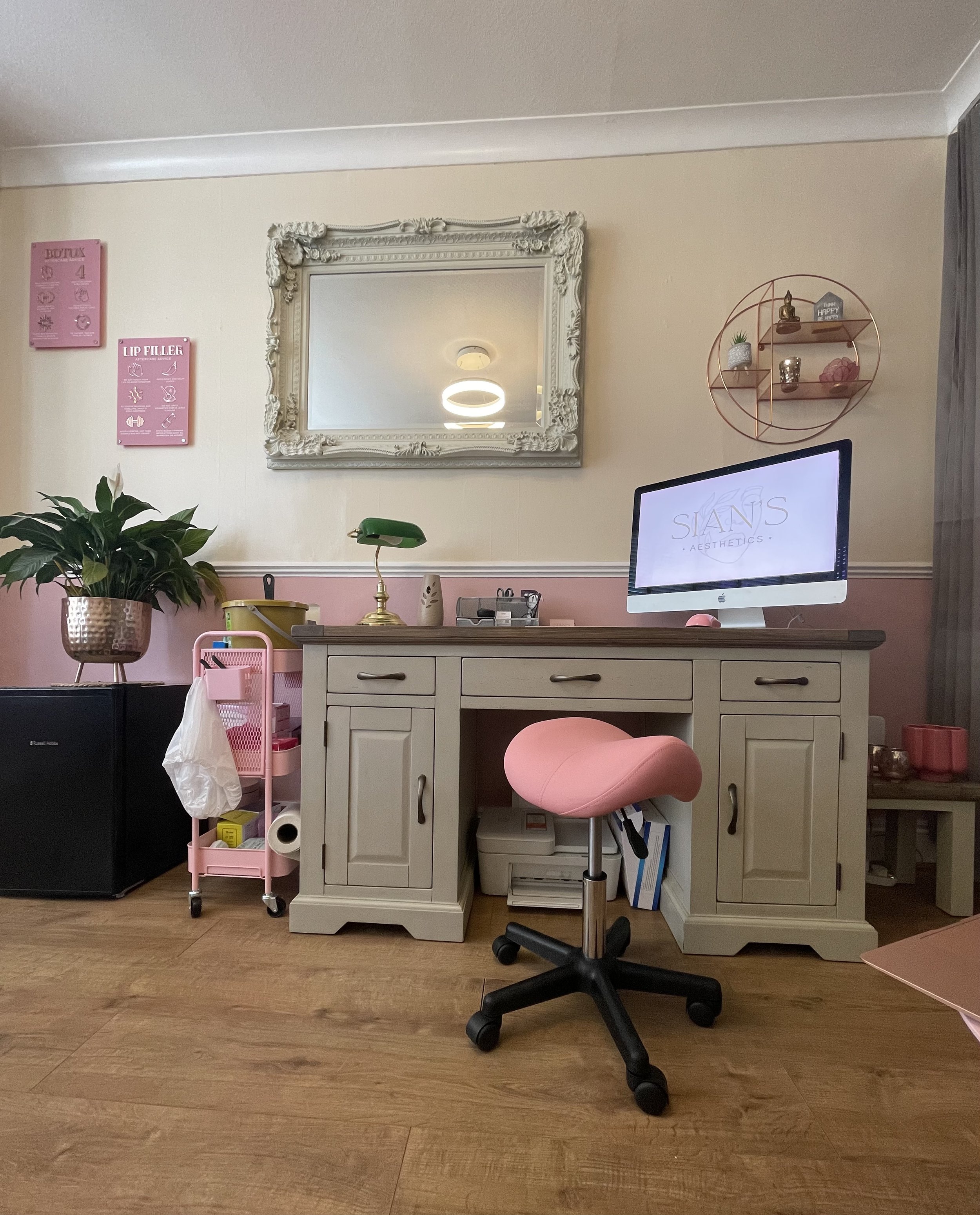 Aesthetic home office setup with a vintage desk, a pink padded stool, and a computer monitor. The wall features a decorative mirror and shelving with small plants and ornaments. A potted plant is on the left, beside a pink rolling cart.