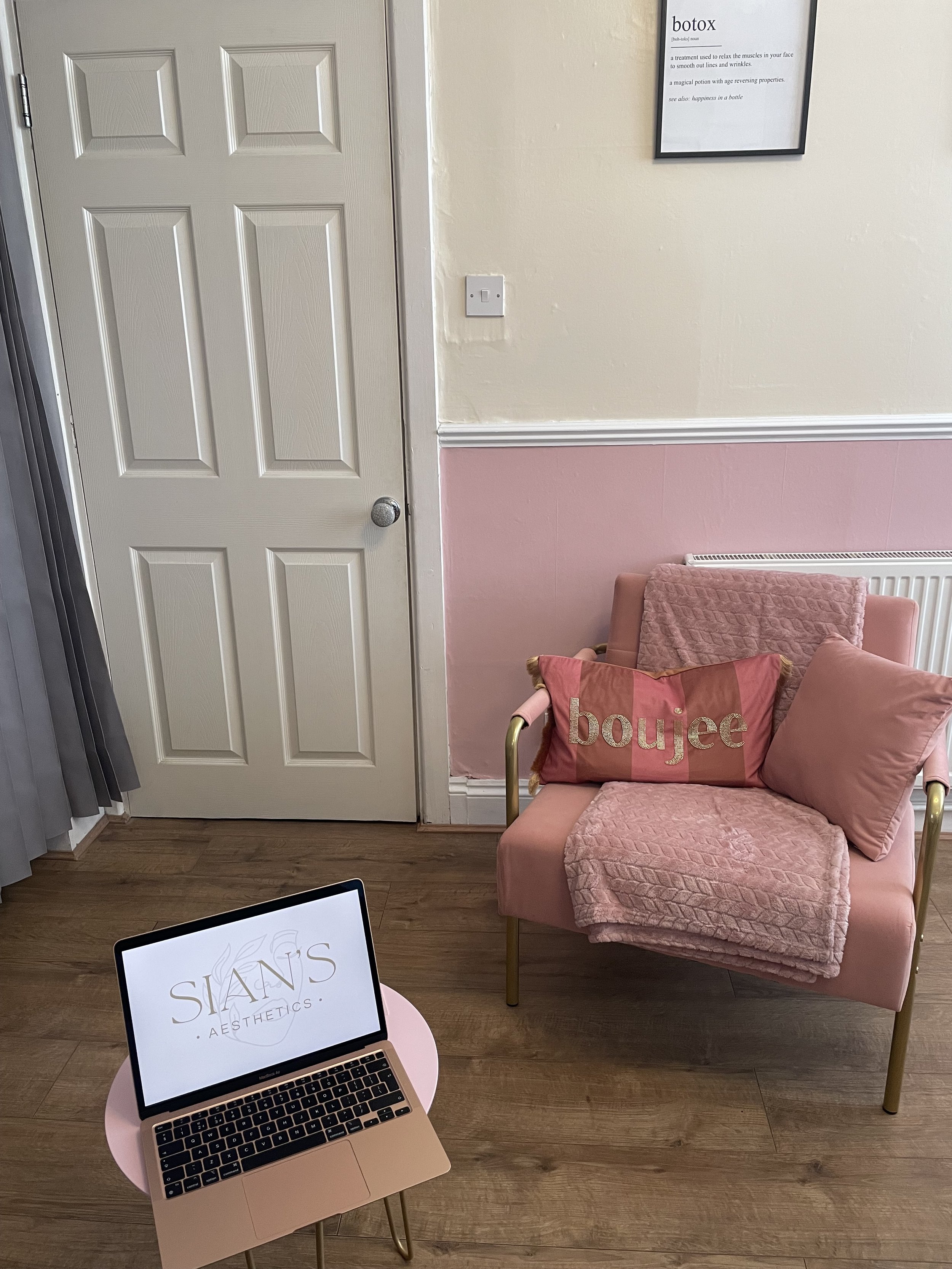 A small room with a pink and cream color scheme, featuring a pink armchair with a "boujee" pillow and a textured blanket. A laptop displaying "SIAN'S AESTHETICS" sits on a round pink side table. A framed definition of "botox" is on the cream wall abo