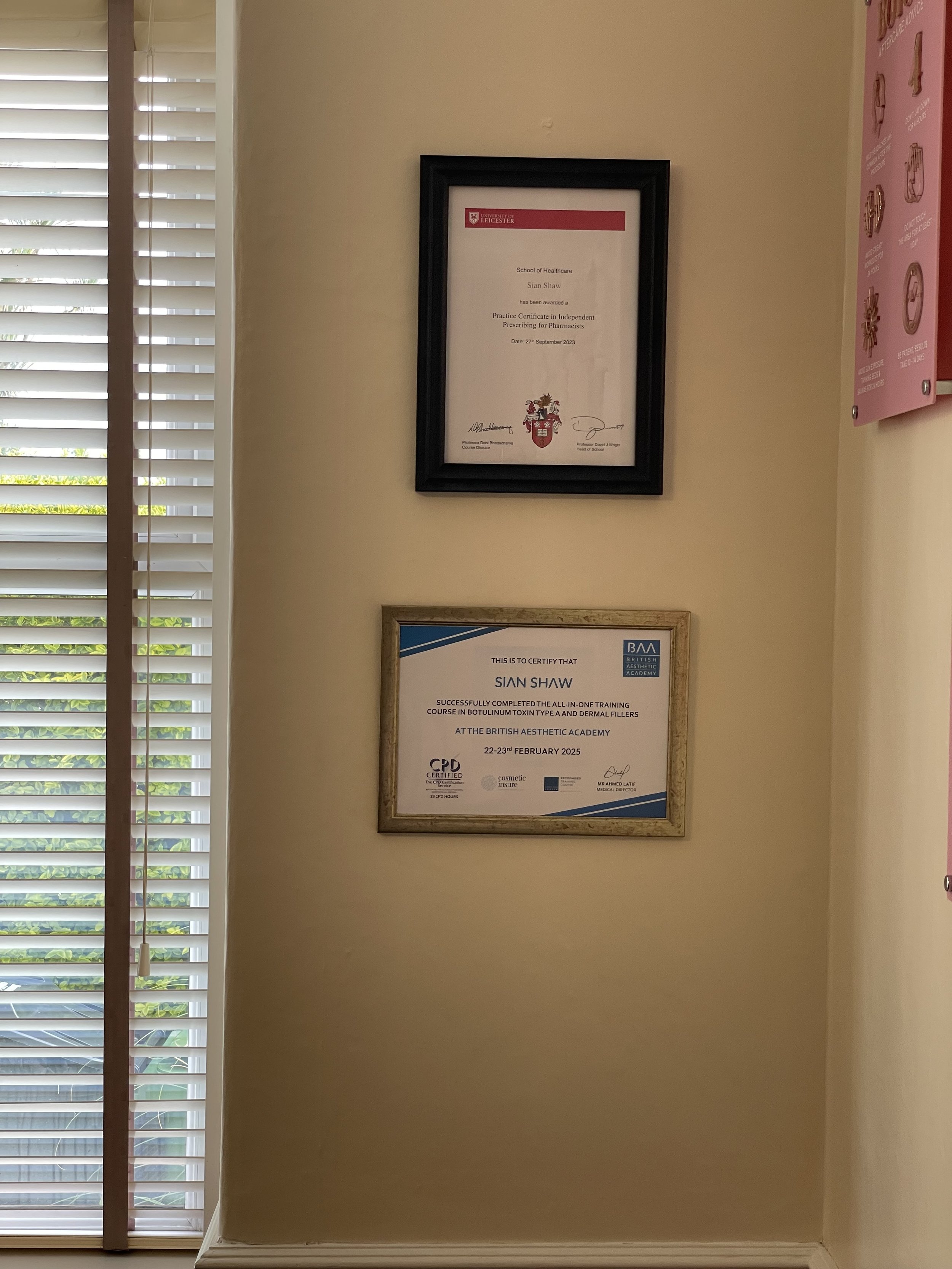 Two certificates hang on a beige wall next to window blinds, one in a black frame and another in a gold frame.