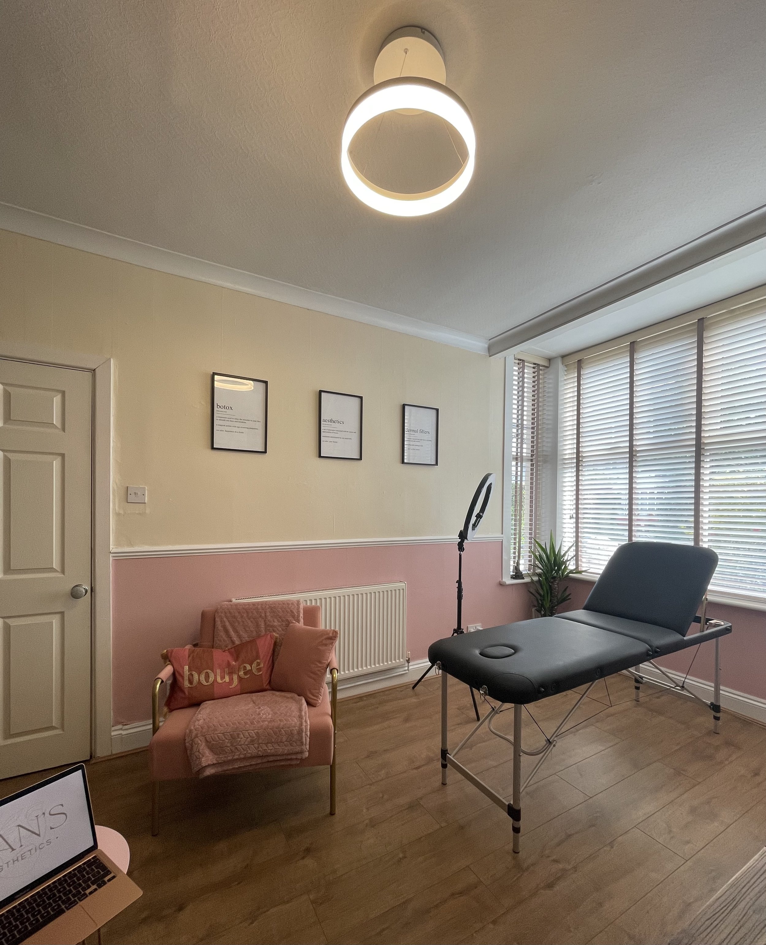 Aesthetic treatment room with a massage table, modern circular ceiling light, pink chair with cushions, framed wall art, and a ring light near a window with blinds. Hardwood floor and potted plant in the corner.