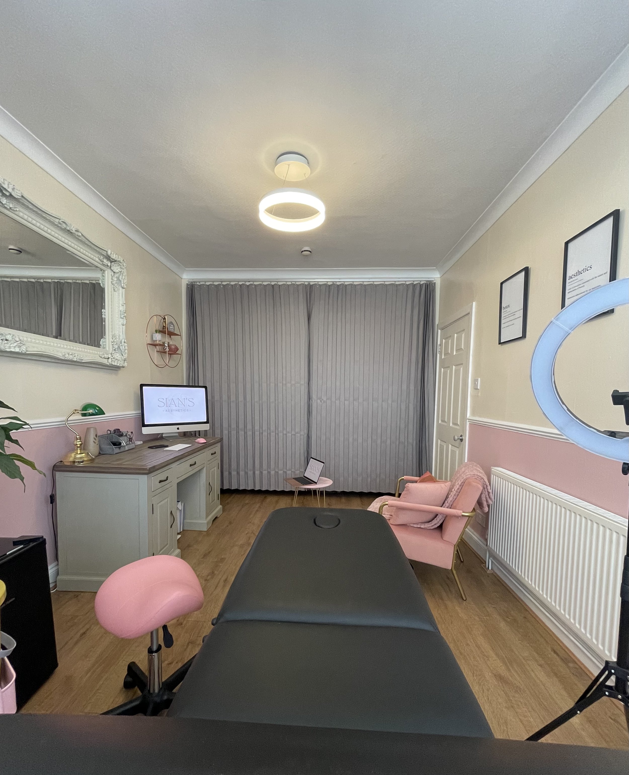Aesthetic treatment room with a comfortable chair, examination table, desk with a computer, and wall-mounted mirror. Pink decor accents include a stool and chair, with a ring light nearby. The room has wooden flooring and curtains covering the window