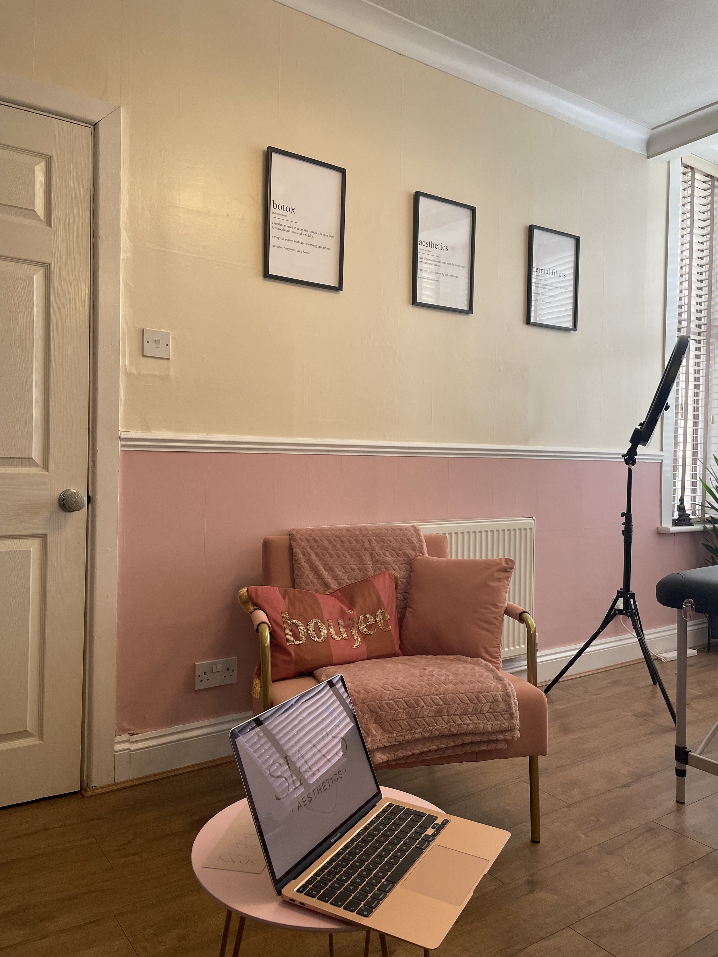 A room with beige and pink walls features three framed motivational posters. There's a pink cushioned chair with a "boujee" pillow and a laptop on a small round pink table. A ring light stand is partially visible to the right, along with a massage or