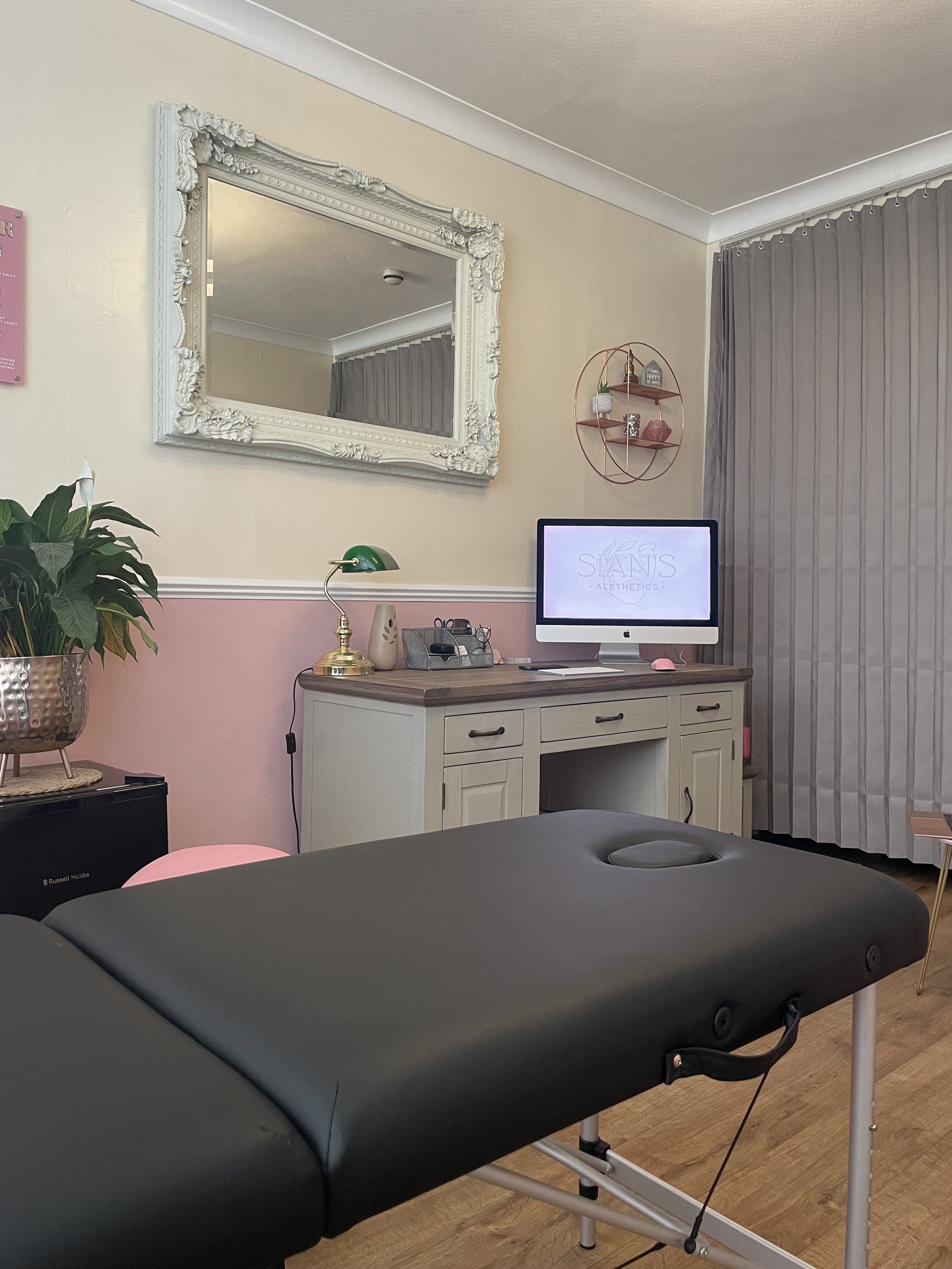 Interior of a beauty or massage salon with a black massage table, a vintage-style white framed mirror on the wall, a desk with a computer, decorative plants, and gray curtains.