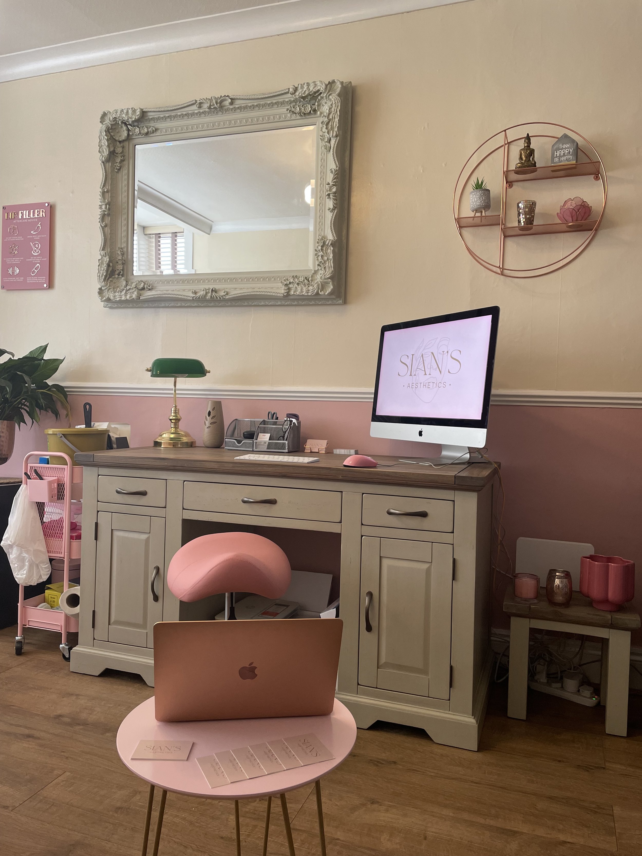 Chic office interior with pink and beige decor. A large ornate mirror hangs above a desk with a computer displaying 'Sian's Aesthetics' logo. A pink chair, laptop, small table with business cards, and decorative items are visible. The room has hardwo