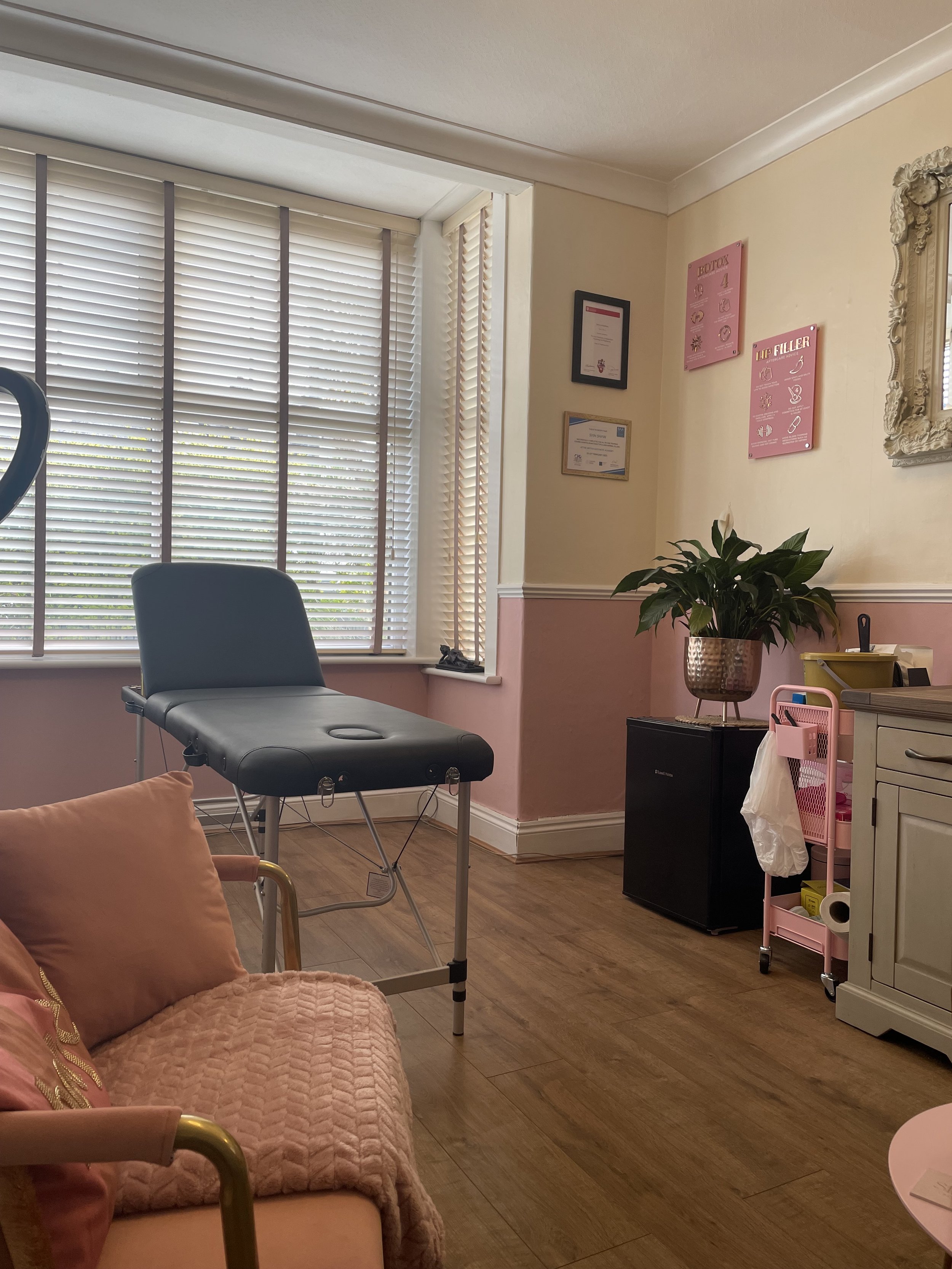 A small, cozy spa or beauty treatment room with a massage table, a pink chair, and various decor. The room has soft pink walls, a window with blinds, framed certificates, and a plant. A cart with supplies and a mirror are also visible.