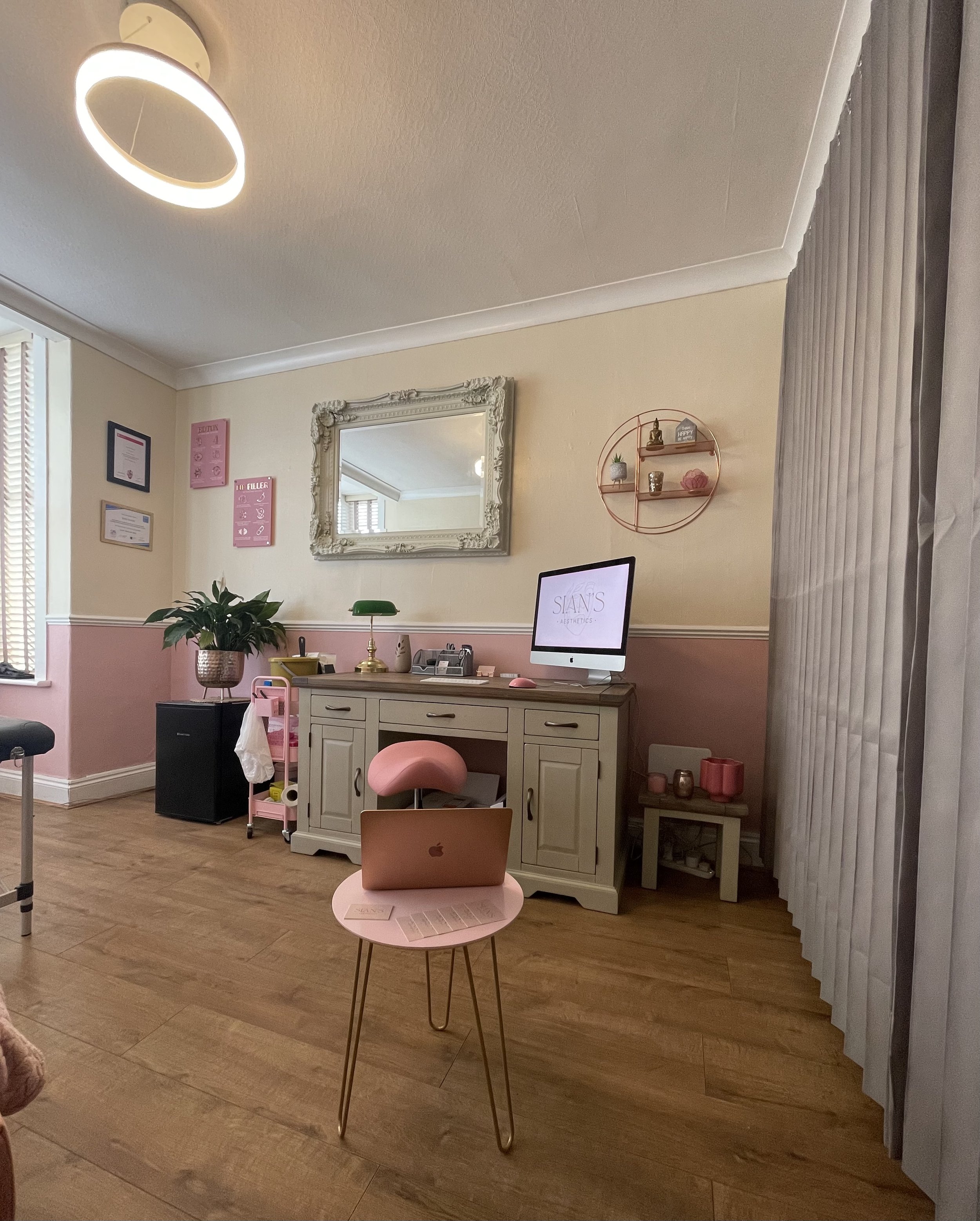 A cozy and stylish home office with pink accents, featuring a wooden floor, a modern ring light, a wall-mounted mirror, a white cabinet displaying a computer screen, and a small pink side table with a laptop placed on it. There's also a potted plant