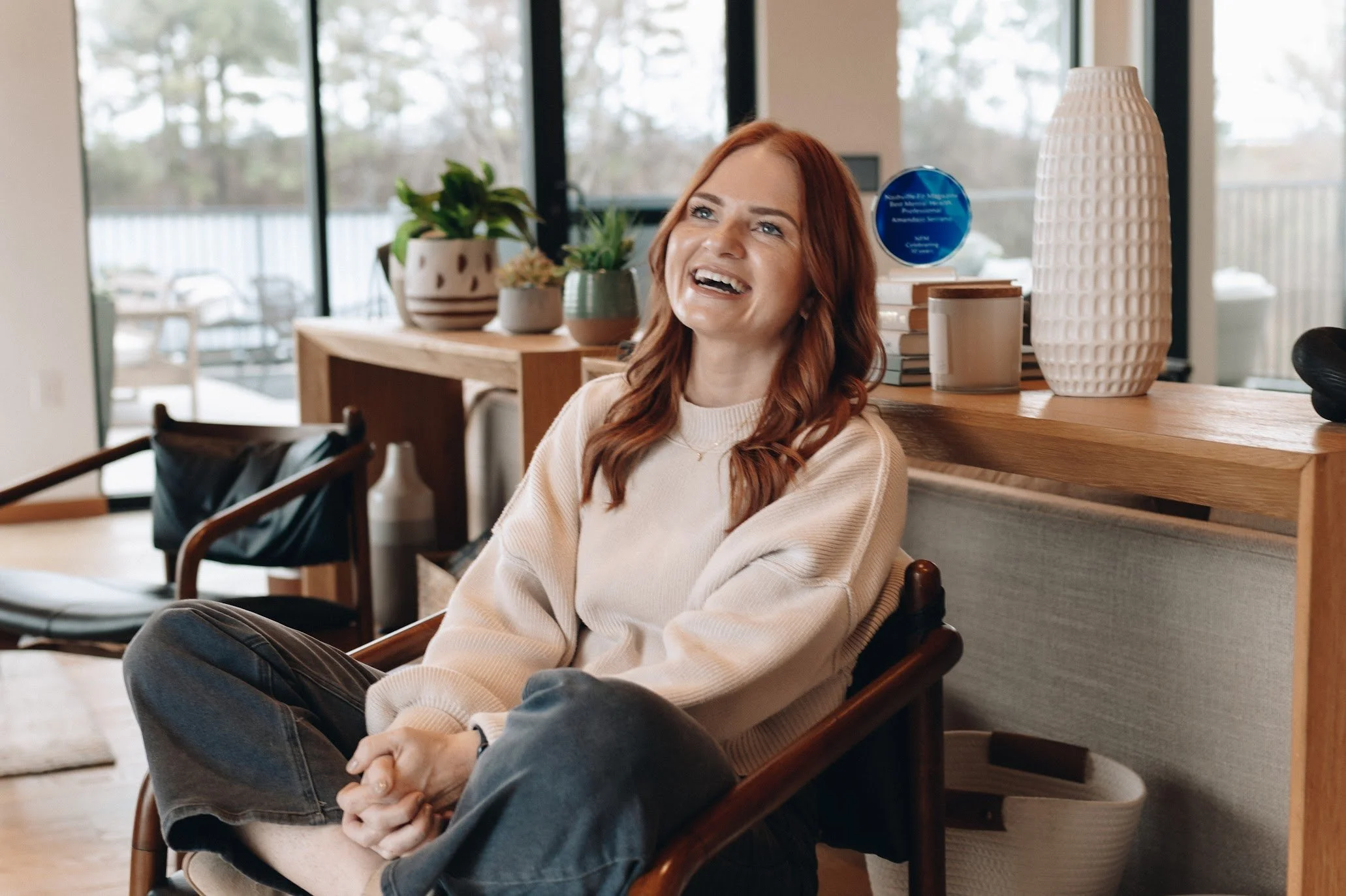 A woman with red hair sitting in a chair, smiling and laughing in a bright room with large windows, potted plants, and decorative objects on a wooden shelf.
