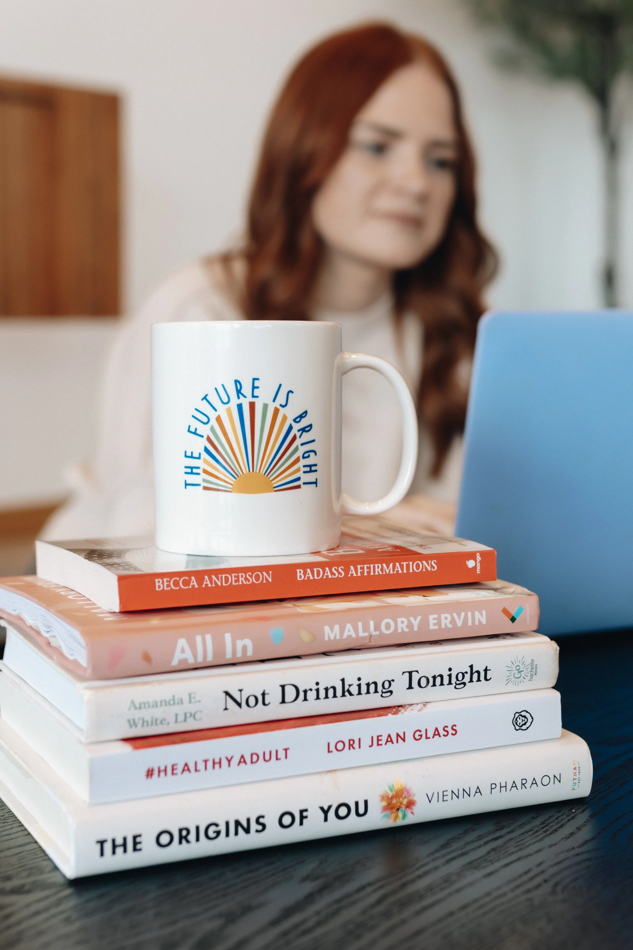 Stack of books on a desk with a white mug on top, featuring a sunrise graphic and the phrase 'The Future Is Bright'. In the background, a woman with red hair is working on a laptop.