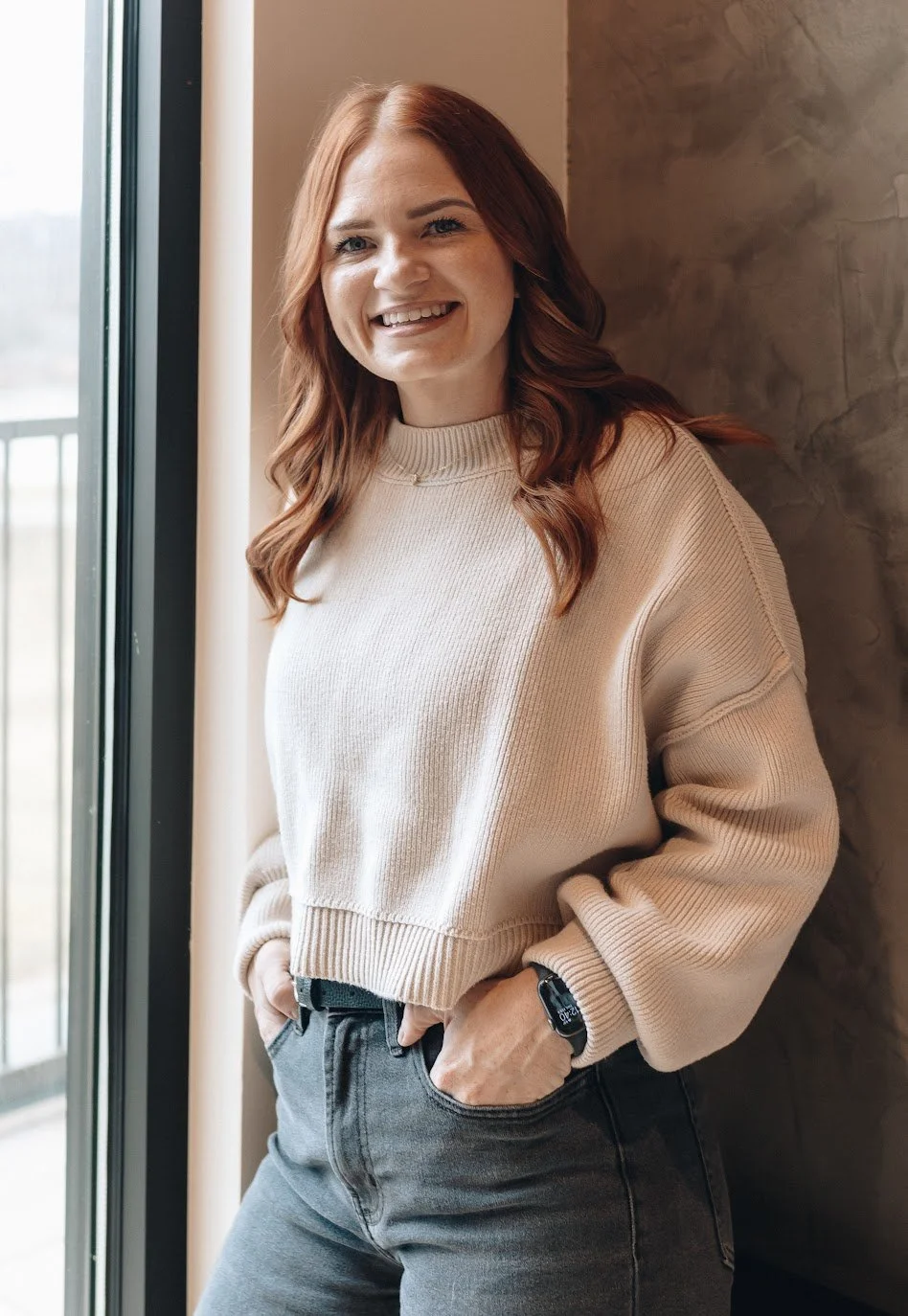 Young woman with red hair smiling, wearing a beige sweater and gray jeans, standing near a window.