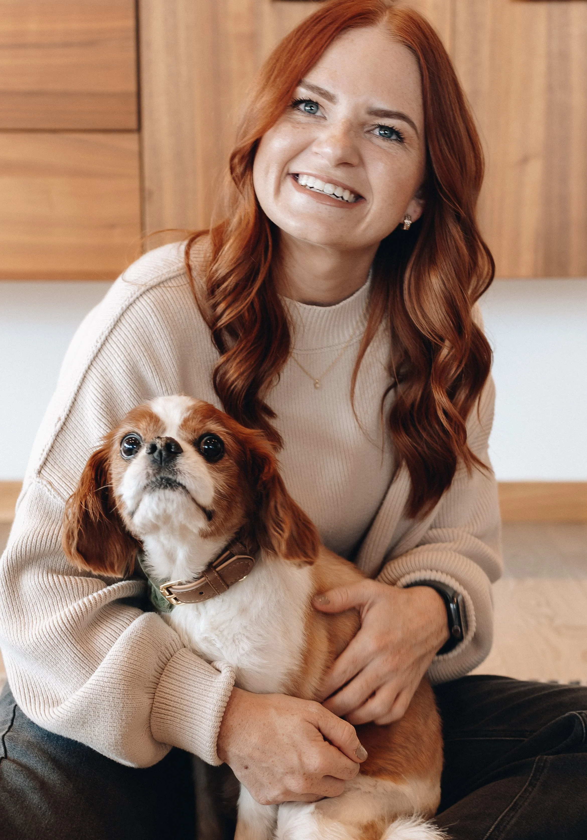 A woman with red hair smiling while holding a small brown and white dog indoors.