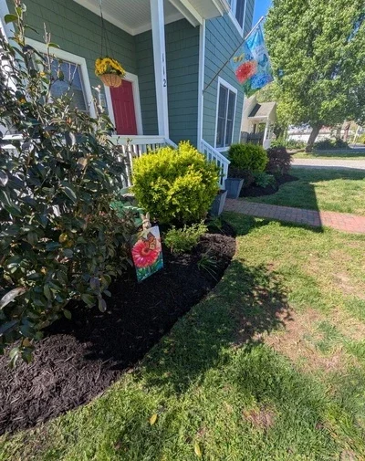 Front yard with green shrubbery, a colorful hanging decoration, and a house with blue siding, white trim, and a red door.