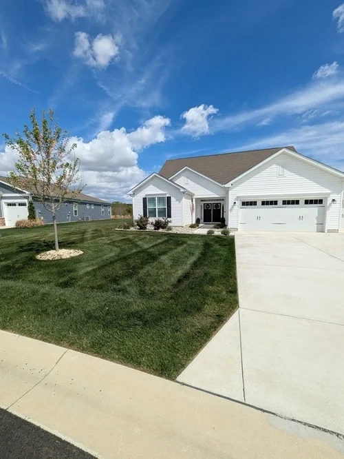 A white house with a gray roof, a front yard with green grass, a small tree, and a driveway leading to a two-car garage. The sky is partly cloudy.