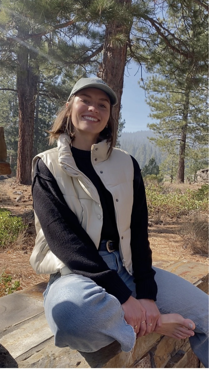 A woman sitting on a log outdoors, smiling at the camera, surrounded by trees and mountain scenery in the background.