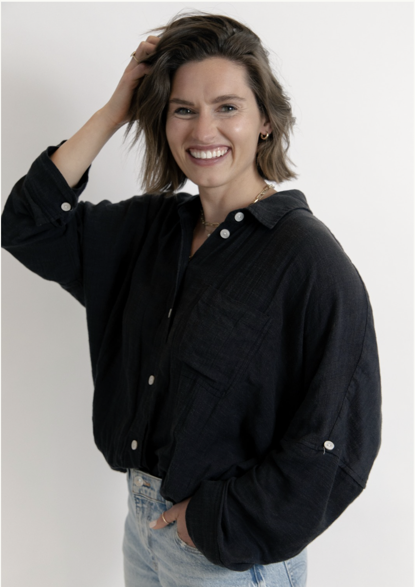A woman with shoulder-length brown hair smiling and touching her hair with her right hand, wearing a black button-up shirt and light blue jeans, standing against a plain white background.