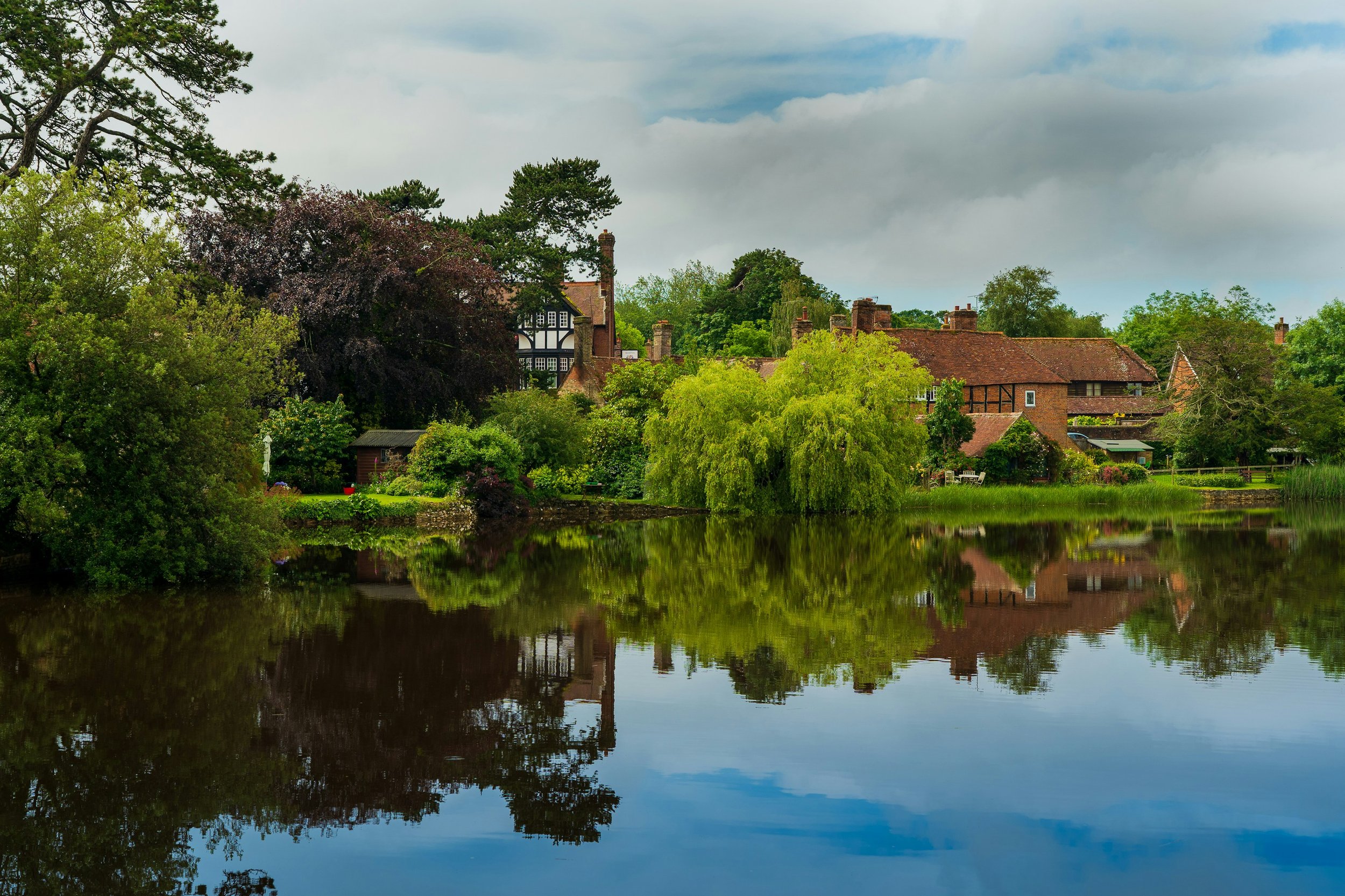 Scenic view of a traditional house by a pond, surrounded by lush greenery and trees, with reflections in the water under a cloudy sky.
