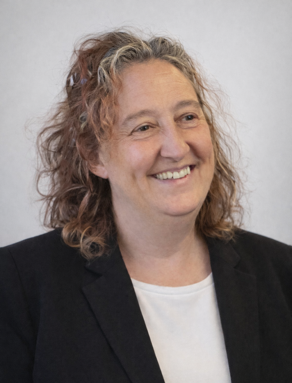 Retrato de una mujer sonriendo, con cabello rizado y cabello rubio con tintes, vestida con saco negro y camiseta blanca, contra fondo gris.