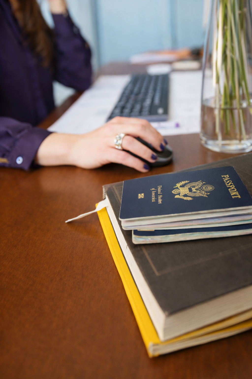 Persona usando una computadora en una oficina, con libros y una visa de pasaporte en primer plano.