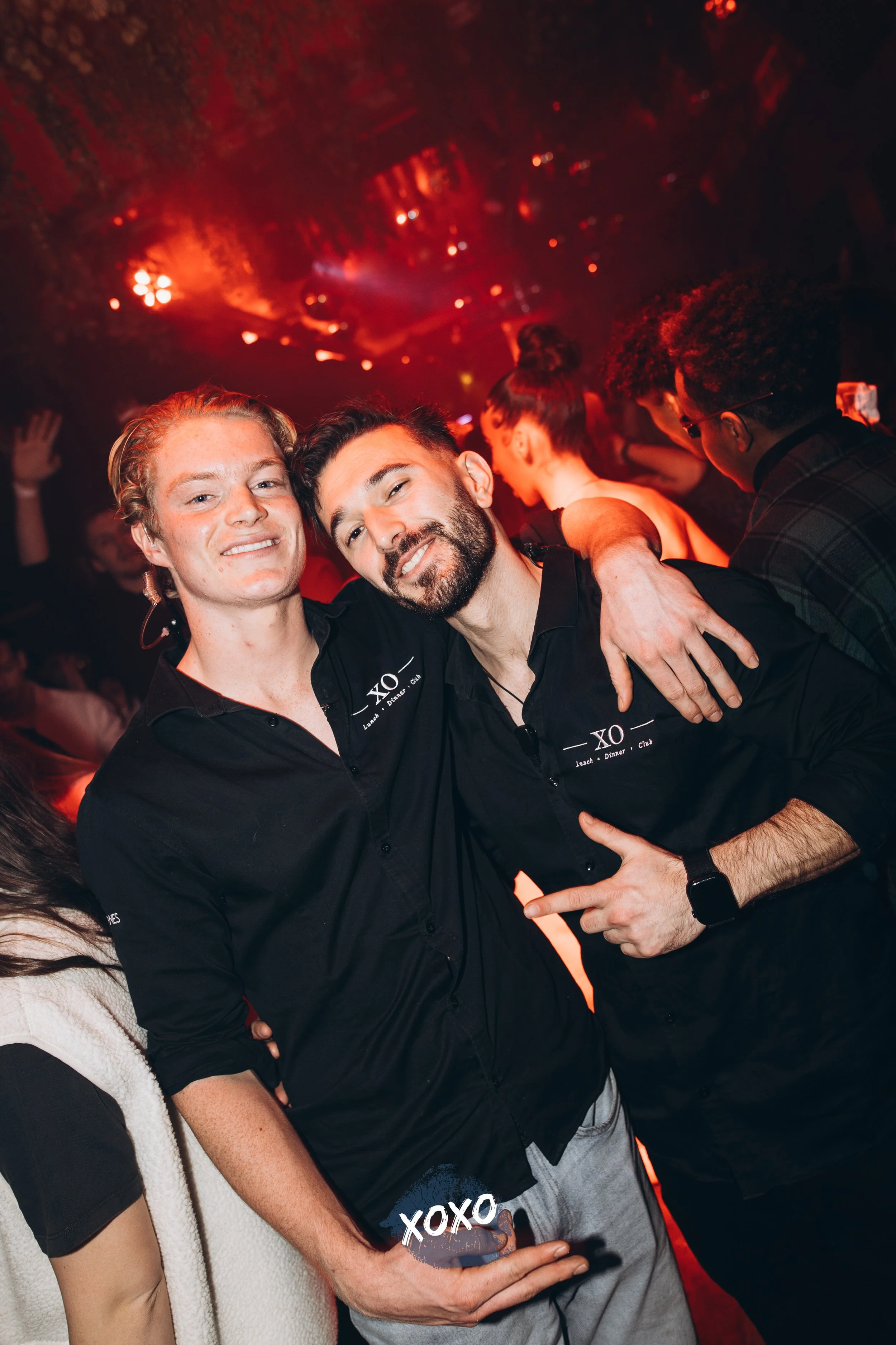 Twee mannen poseren in een club met rode verlichting, omringd door andere mensen en feestverlichting.