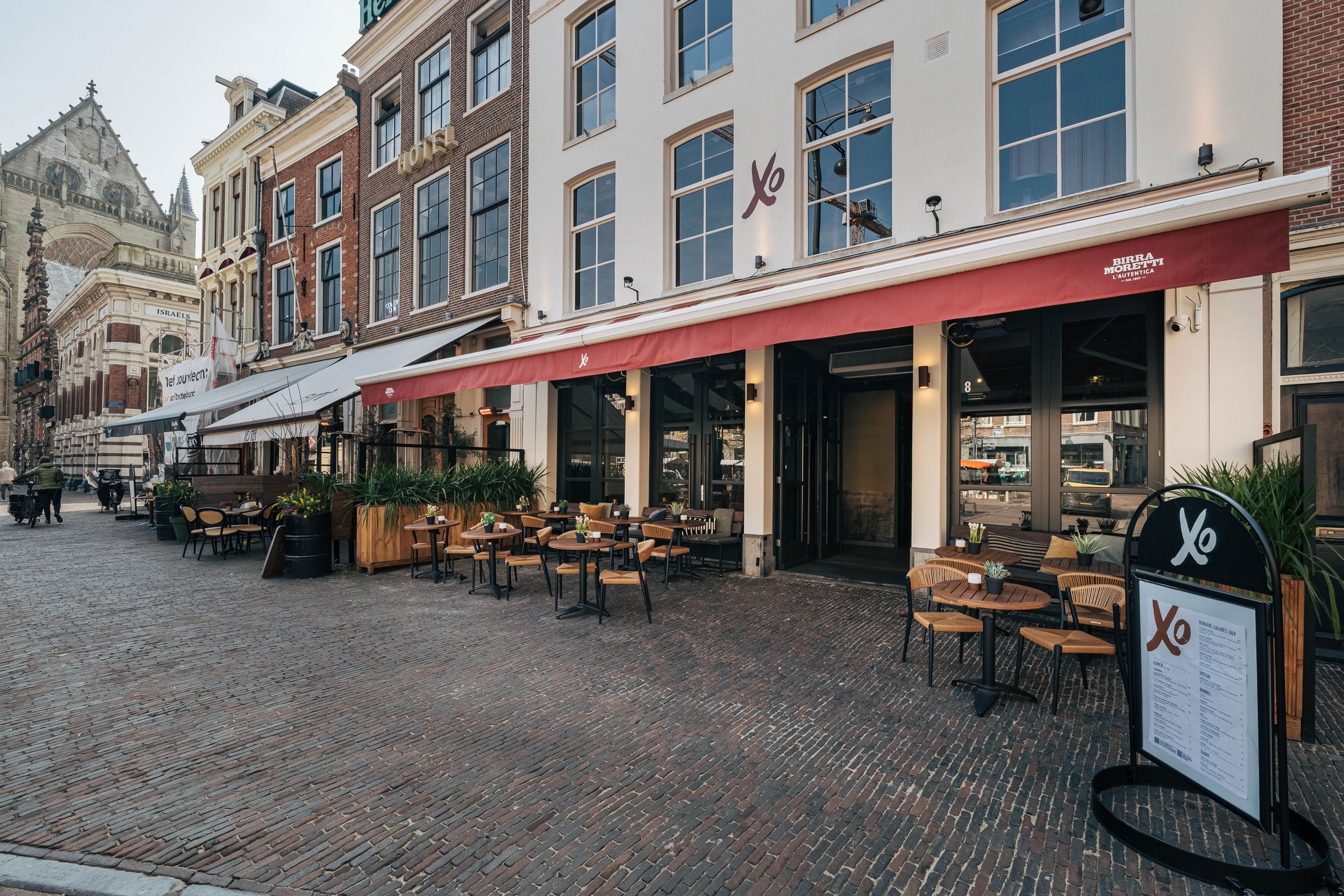 Terras van een restaurant op een geplaveid plein met enkele tafels en stoelen, omgeven door historische gebouwen met een kerk in de achtergrond.