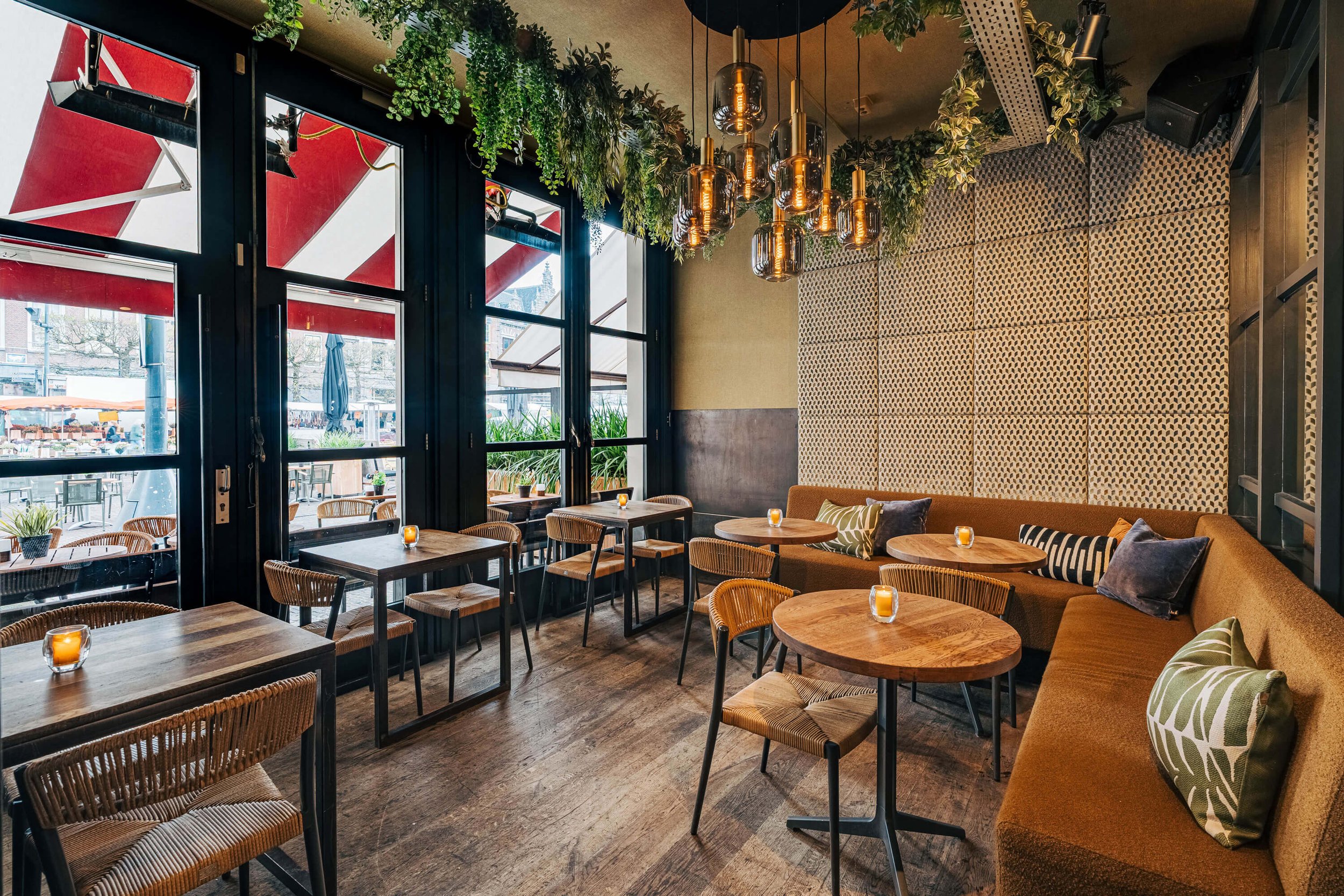 Een stijlvol ingerichte café-interieur met houten tafels, stoelen en banken. Er zijn sierlijke hanglampen aan het plafond en groene planten boven de ramen. De ruimte biedt uitzicht op een terras met rode zonneschermen.