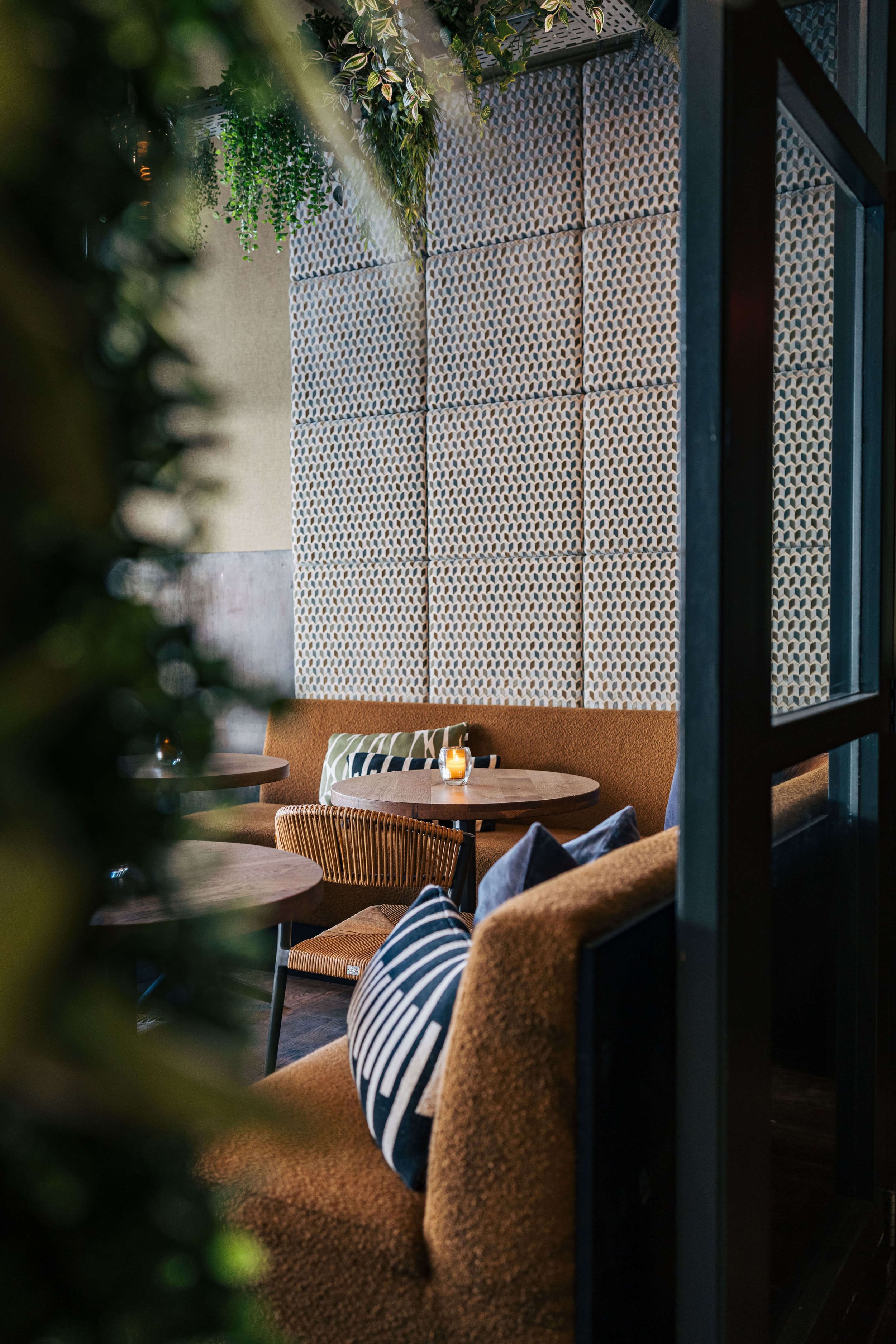 Gezellige café interieuropname met bruine banken, ronde houten tafels, kussens en groene planten aan het plafond, naast een decoratieve muur.
