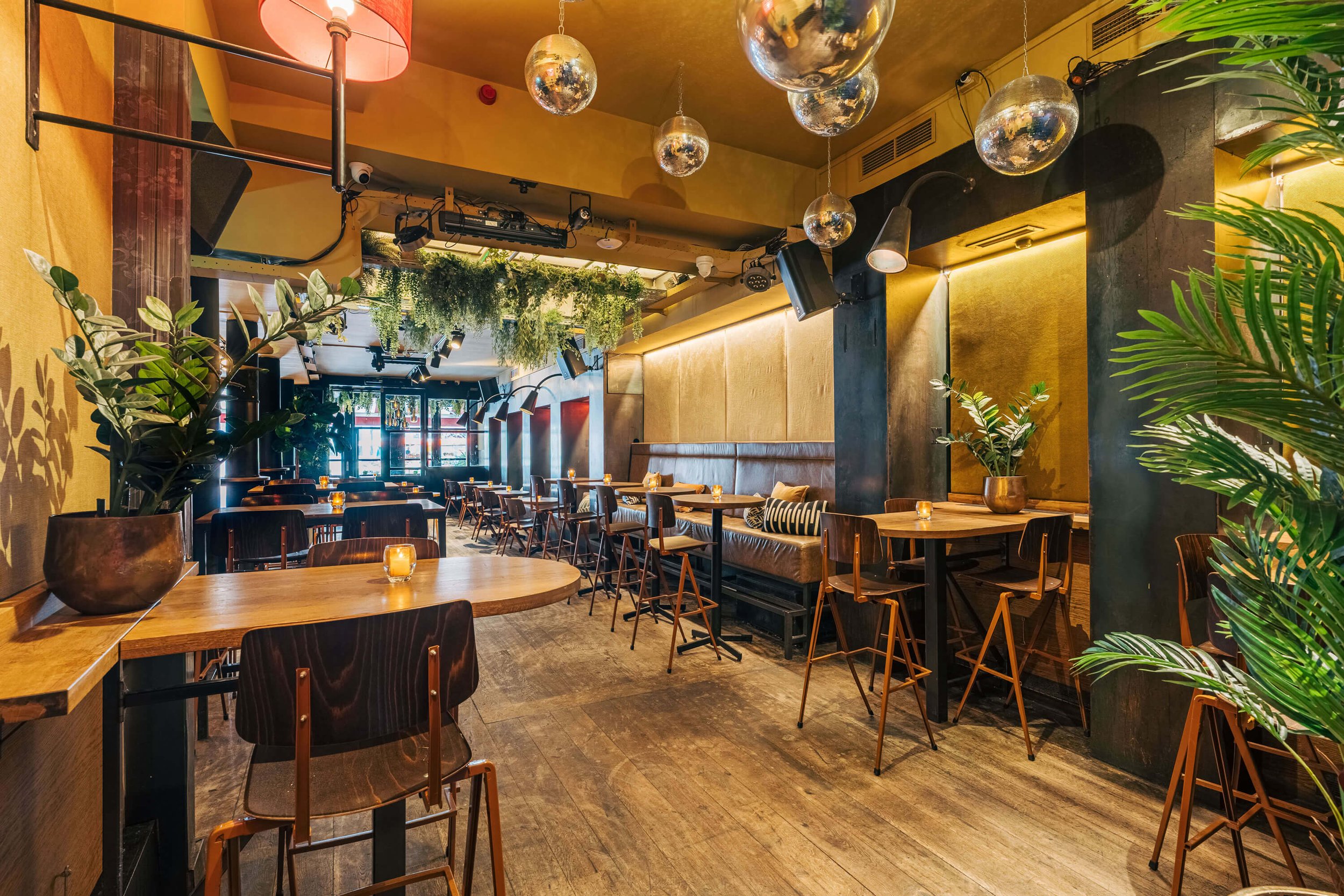 Interieur van een stijlvolle café met houten tafels en stoelen, sfeerverlichting, groene planten en moderne decoratie, inclusief hangende lampen en spiegels aan het plafond.