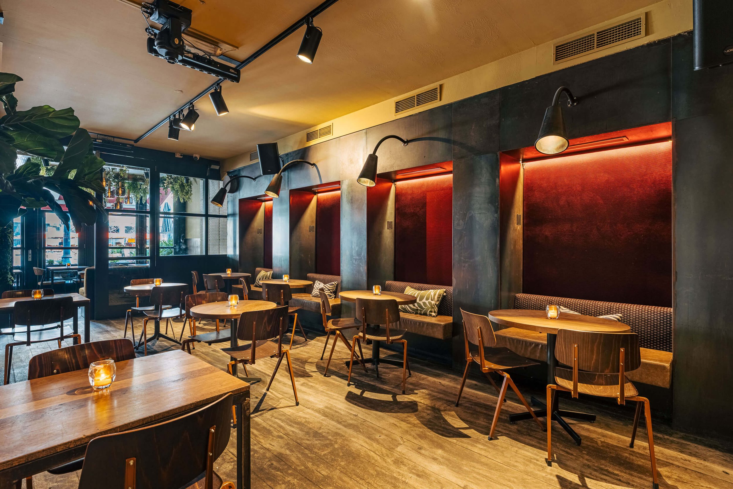 Interieur van een moderne, sfeervol verlichte bar met houten tafels en stoelen, pluchen banken aan de zijkant en grote ramen met uitzicht naar buiten.