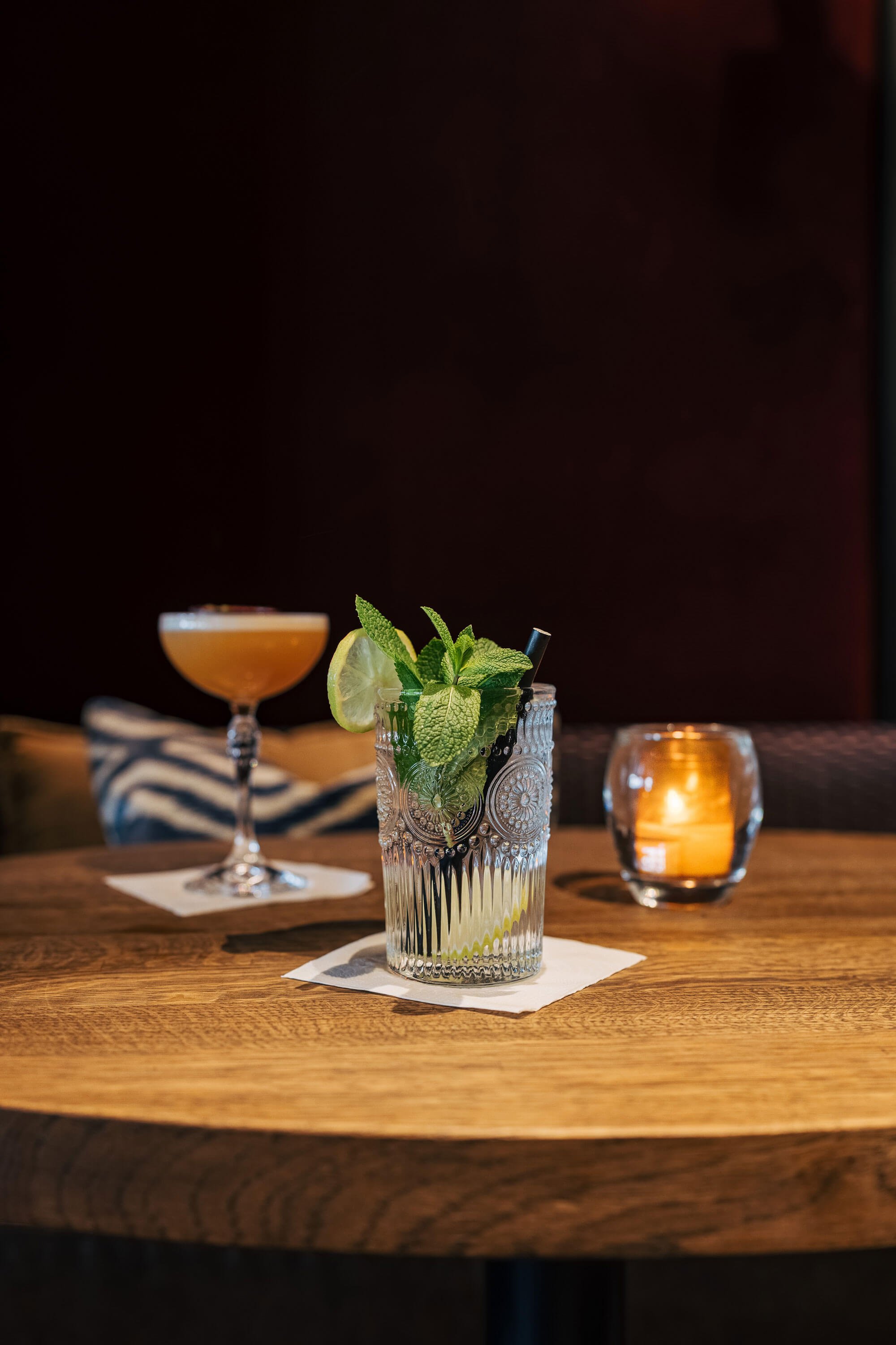Twee cocktails op een houten tafel, een met munt en limoen in een glazen beker en een oranje cocktail in een coupeglas. Er staat ook een kaars op tafel.