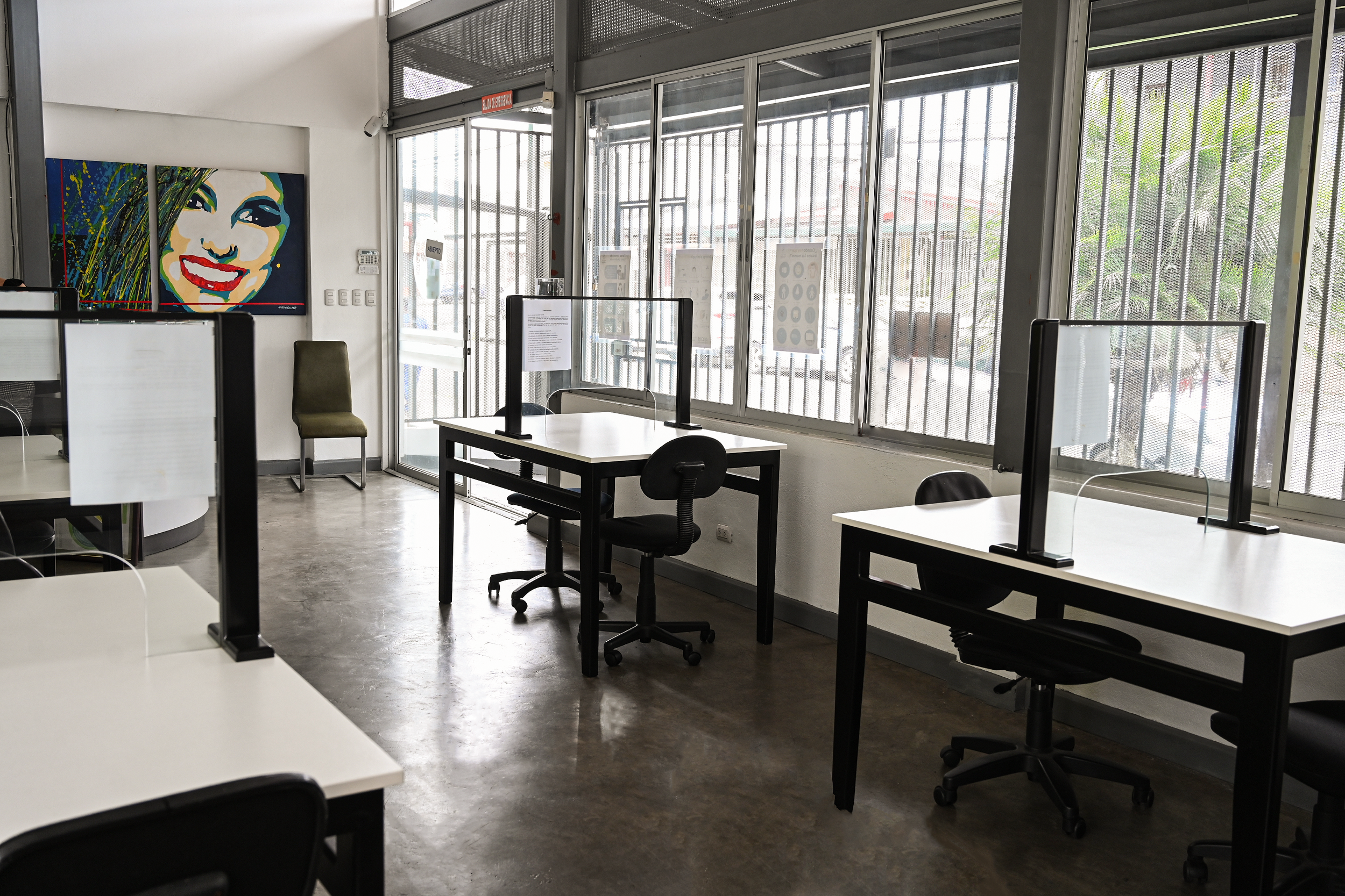 Interior of an office with desks and chairs separated by glass partitions, a colorful abstract portrait on the wall, large windows with bars, and an exit sign.