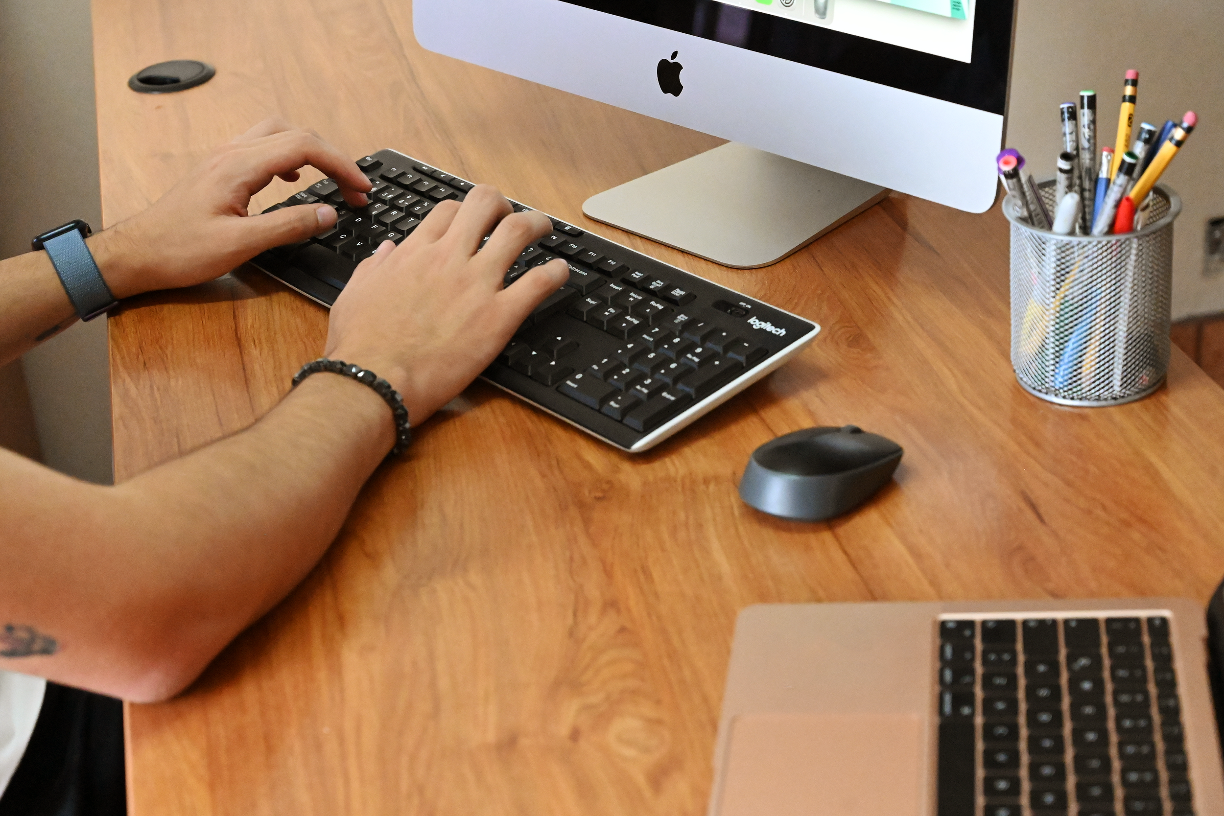 Person typing on Logitech keyboard at wooden desk with Apple iMac, wireless mouse, and pencil holder.